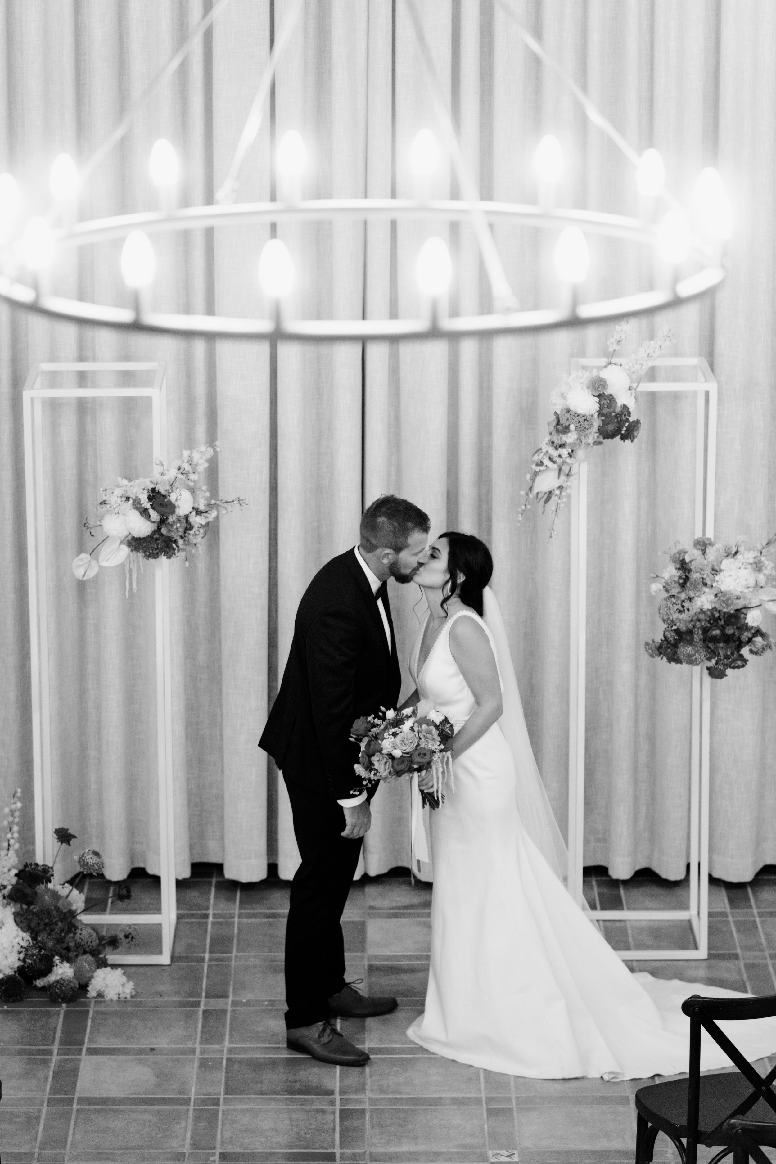 A black and white photo of a bride and groom sharing a kiss during their wedding ceremony, with floral arrangements and a decorative backdrop.