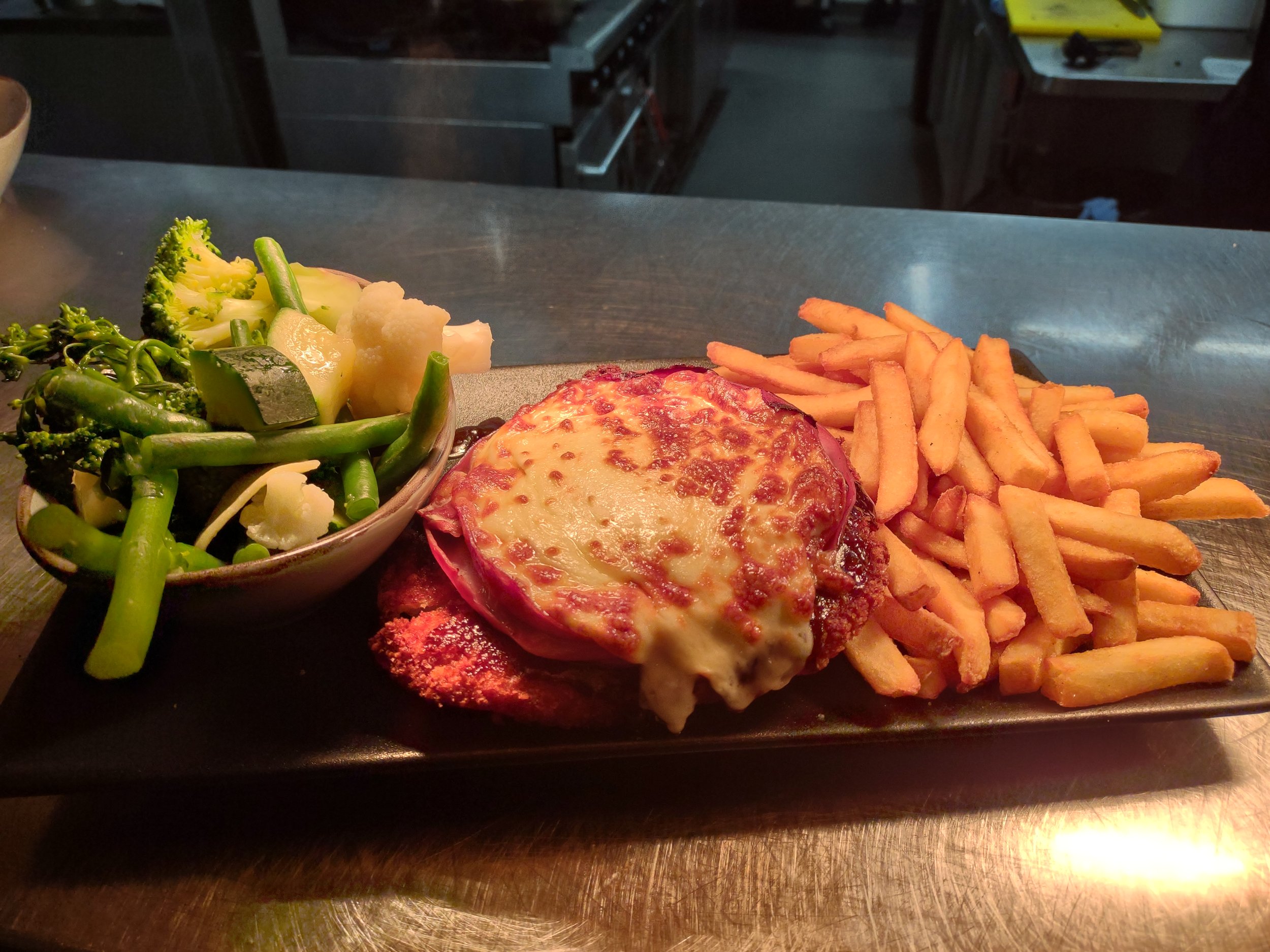 A black rectangular plate with a serving of steamed broccoli, green beans, cauliflower, a breaded meat cutlet with melted cheese on top, and a side of French fries.