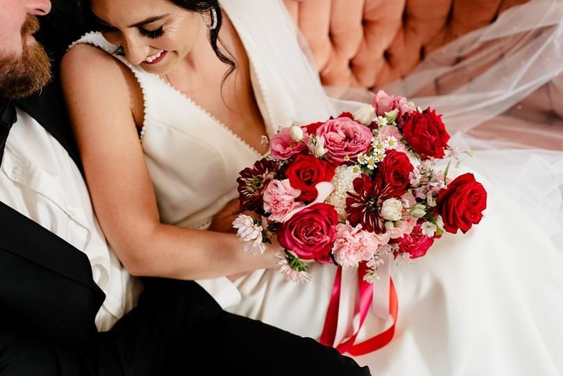 Bride holding a colorful bouquet of red, pink, and white flowers, smiling at her partner during a wedding ceremony.