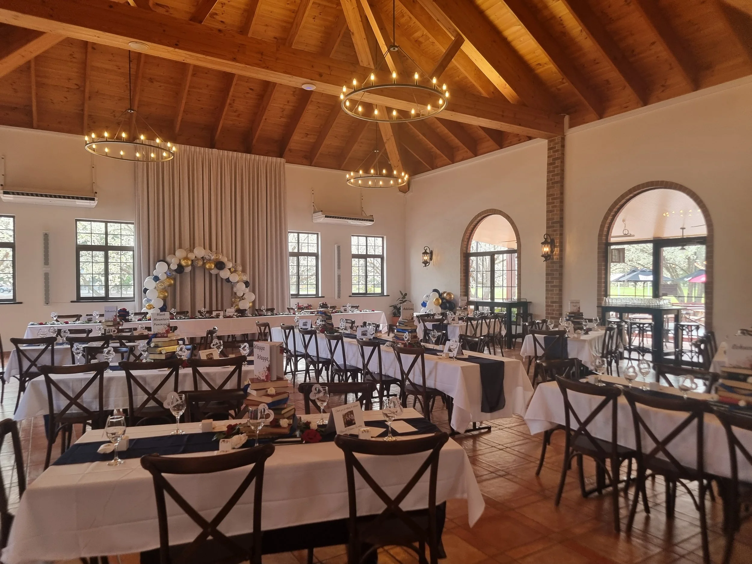 Elegant event space with tables set for a celebration, arranged with white tablecloths, black table runners, glasses, and decorated with books, balloons, and signs, under a wooden vaulted ceiling with circular chandeliers.