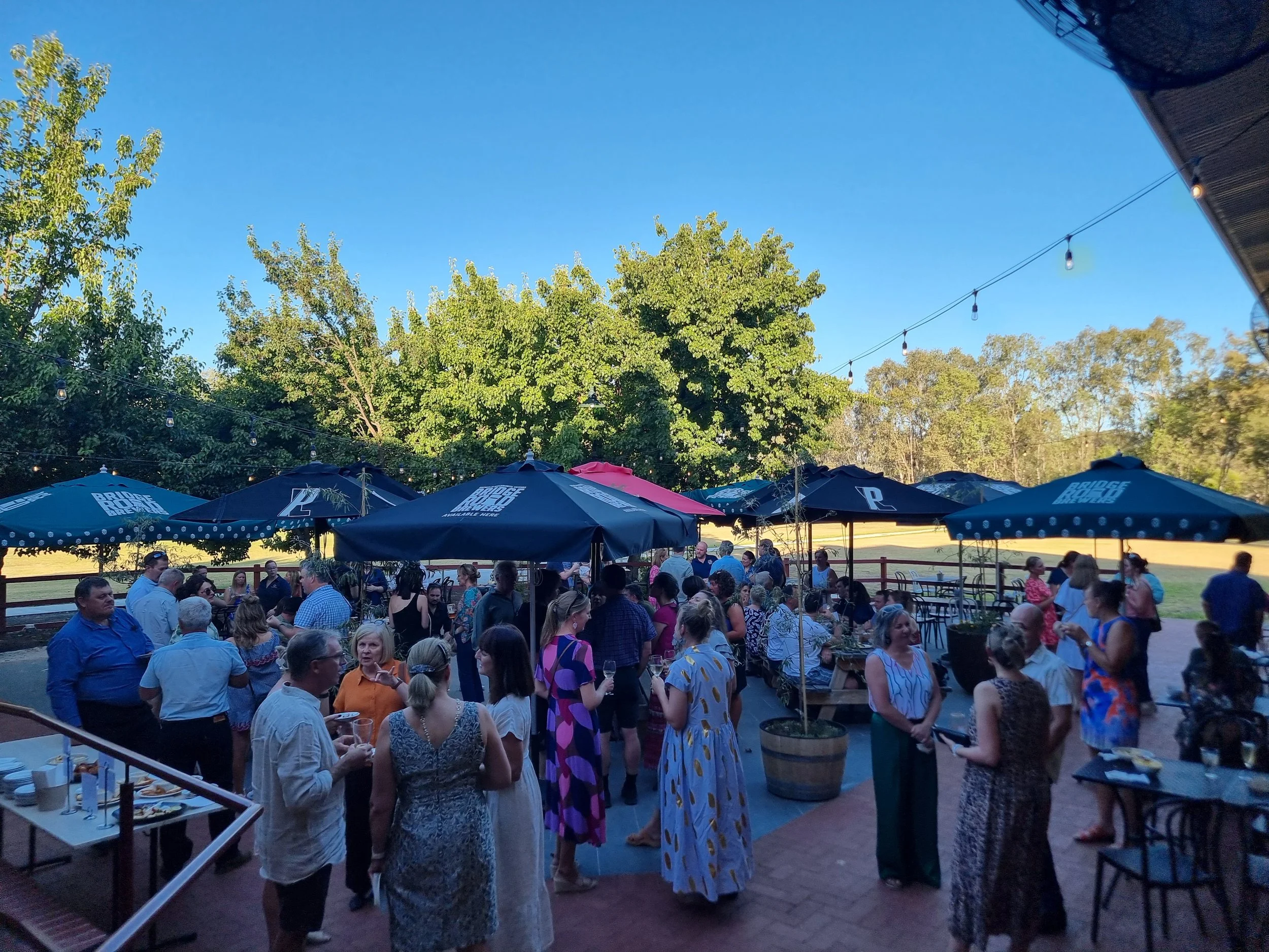 Outdoor social gathering with people mingling under patio umbrellas against a backdrop of trees and a clear blue sky.