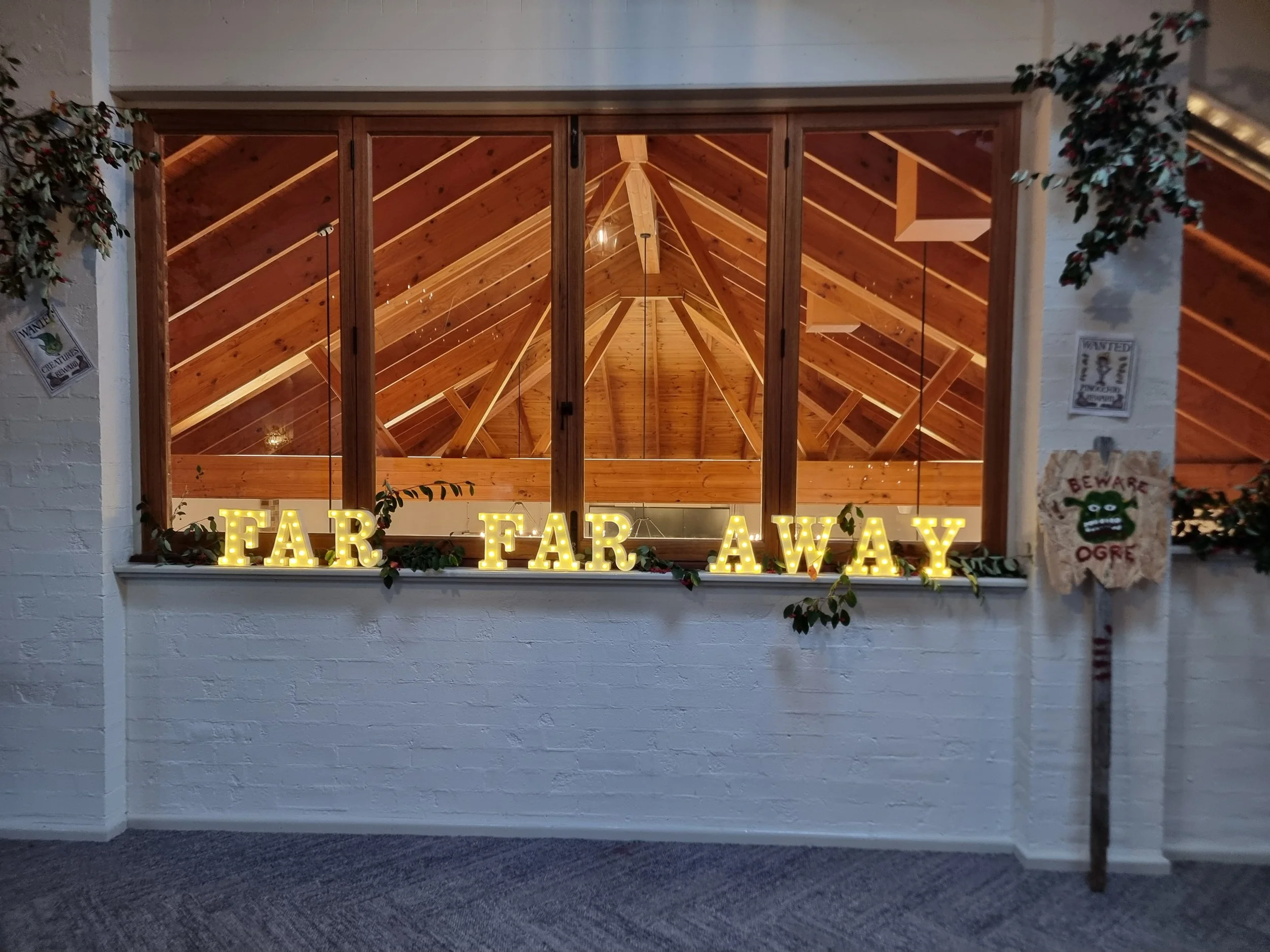 Decorative sign on windowsill spelling 'FAR FAR AWAY' with illuminated gold letters, floral decorations, and a white brick wall, overlooking a wooden attic with exposed beams.