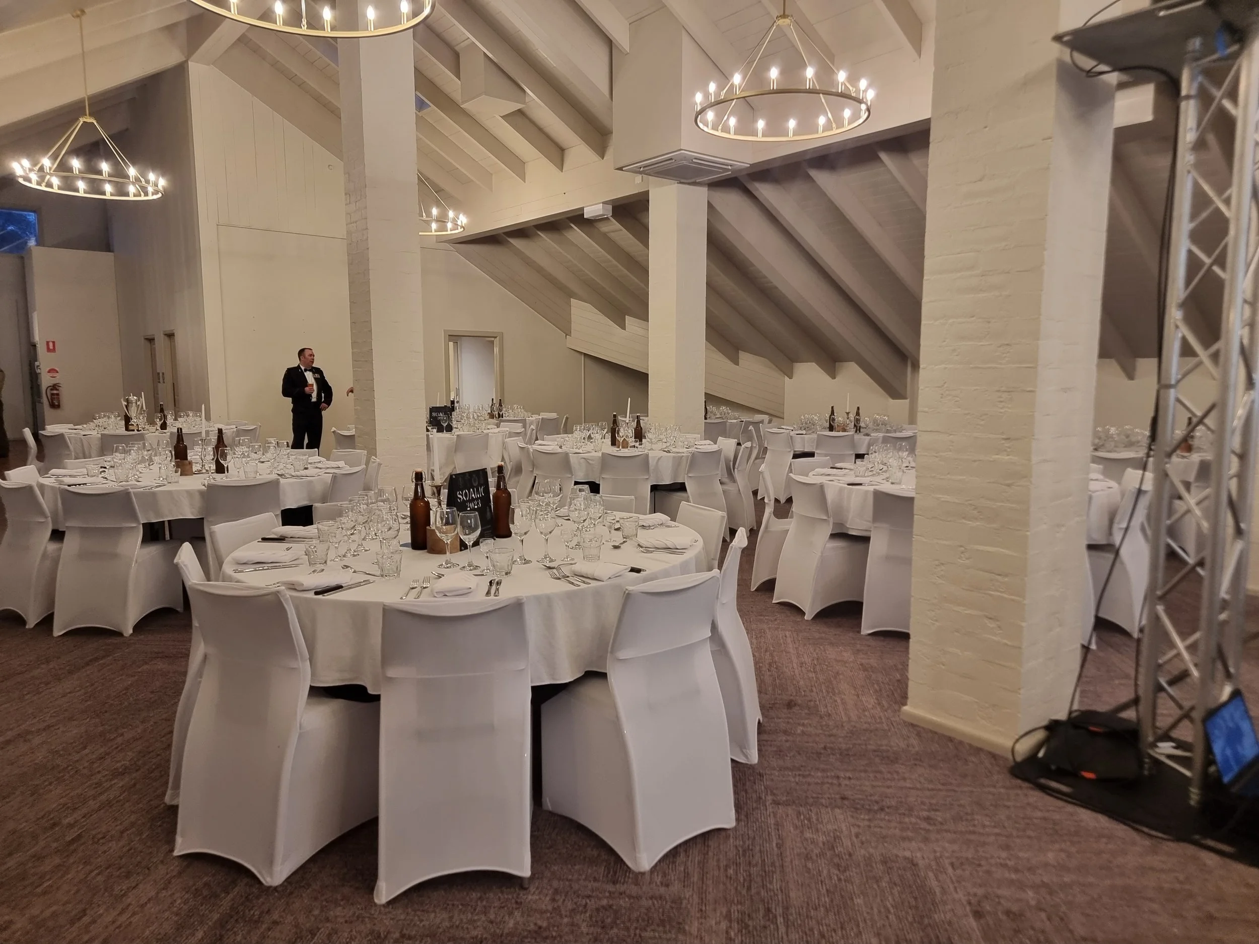 A banquet hall set up with round tables covered with white tablecloths, each surrounded by white chairs with slipcovers, and decorated with glasses, silverware, and bottles, ready for an event.