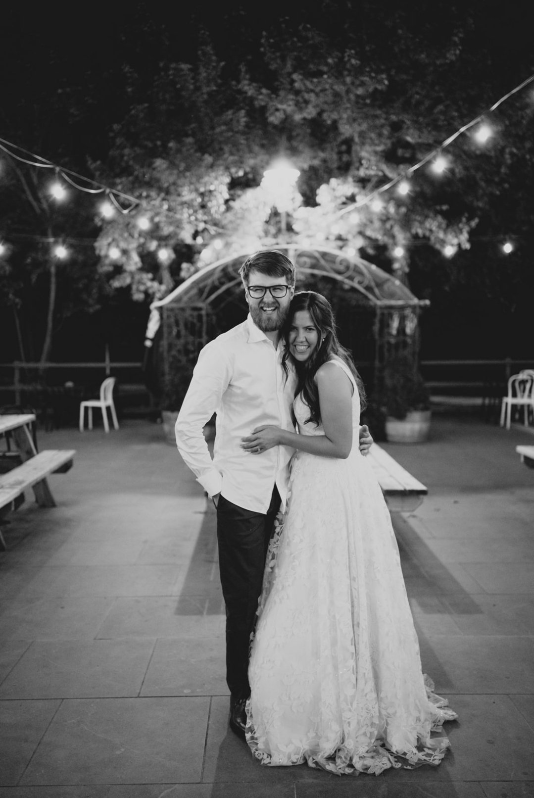 A black-and-white photo of a smiling couple, dressed in wedding attire, standing close together outdoors at night with string lights overhead.