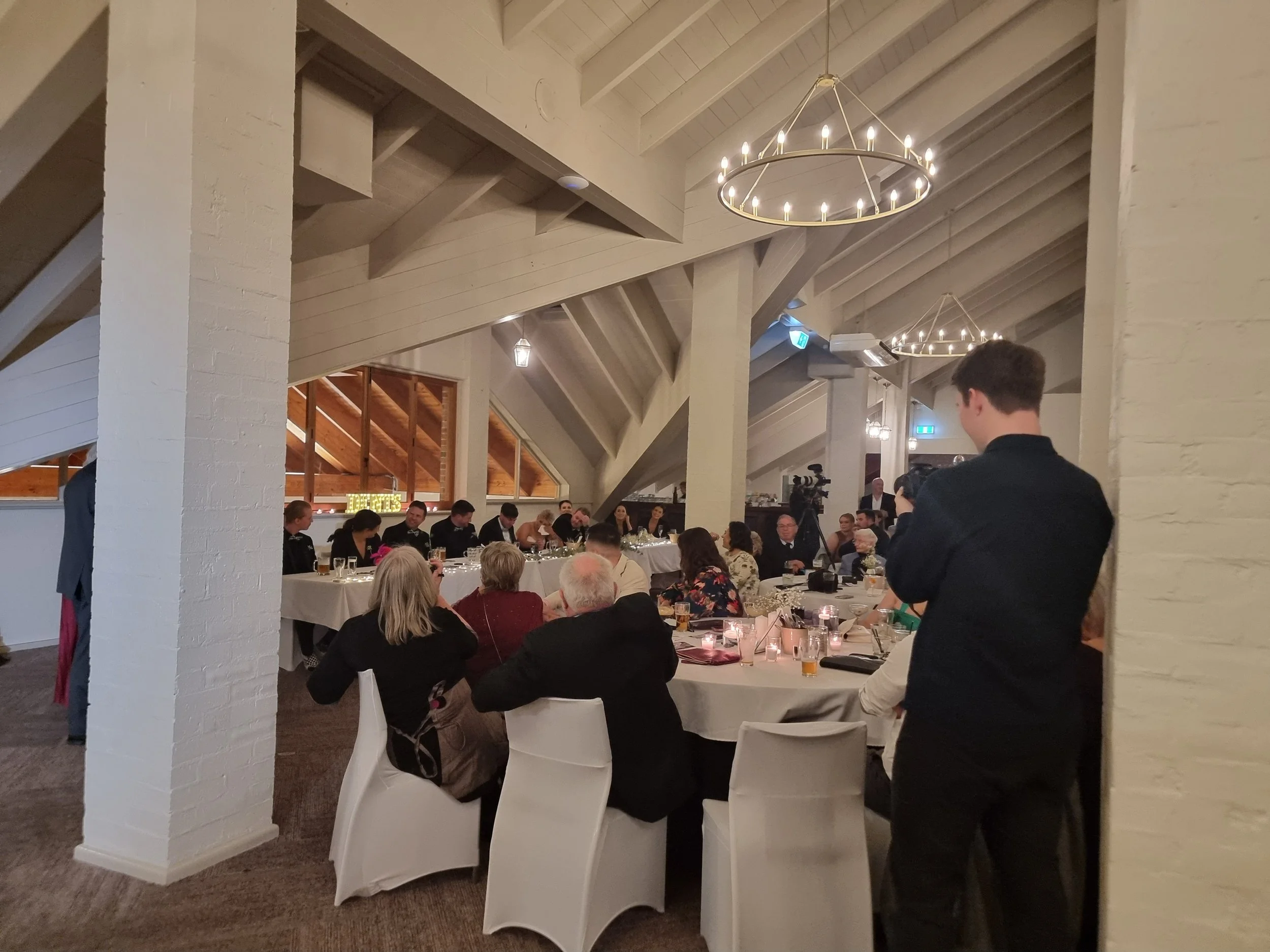 A formal event in a decorated banquet hall with round tables, white chairs, and chandeliers, where guests are seated and a person is filming.