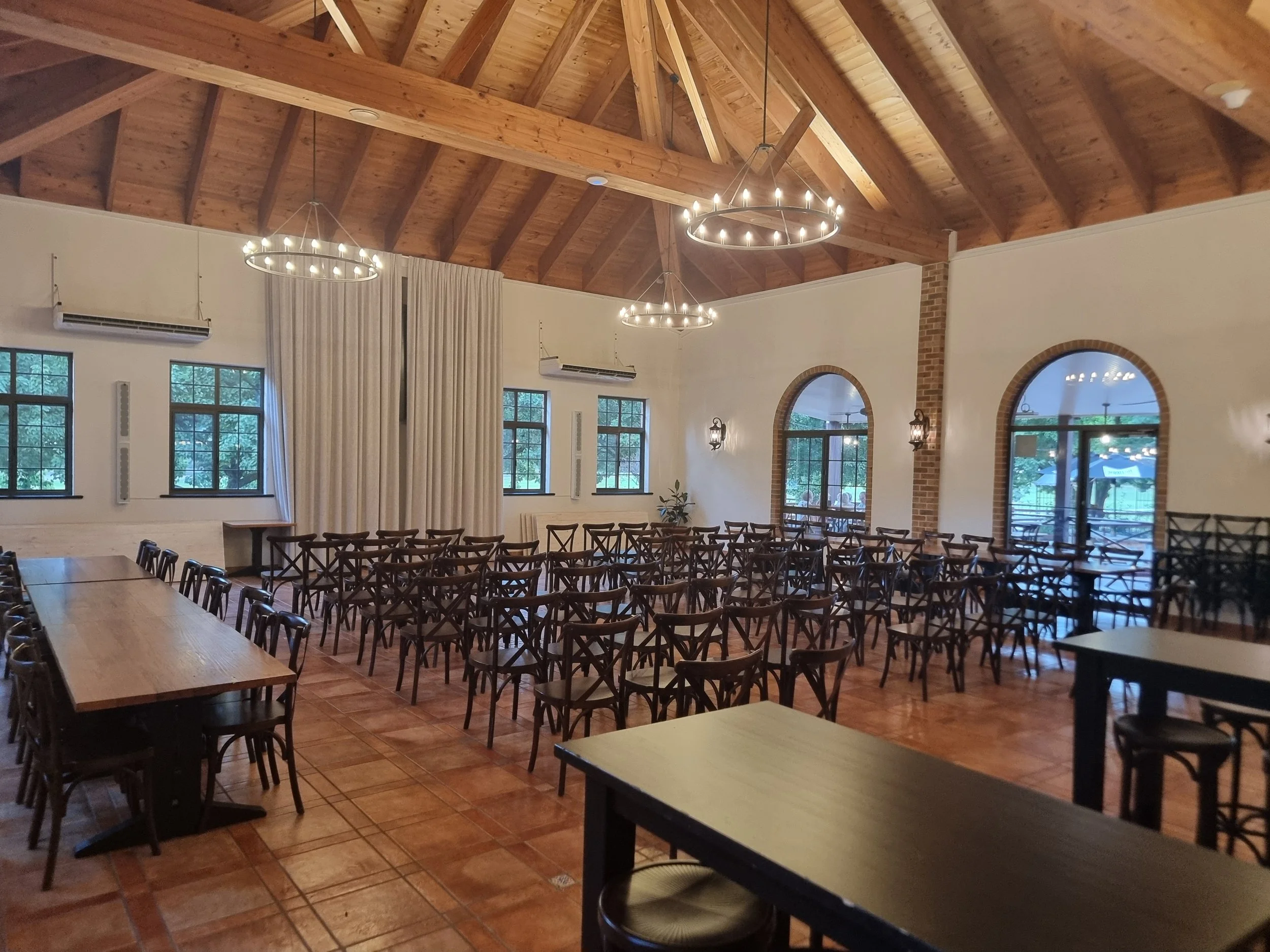 Empty event hall with wooden ceiling, multiple rows of wooden chairs, large windows, brick arches, and chandeliers.