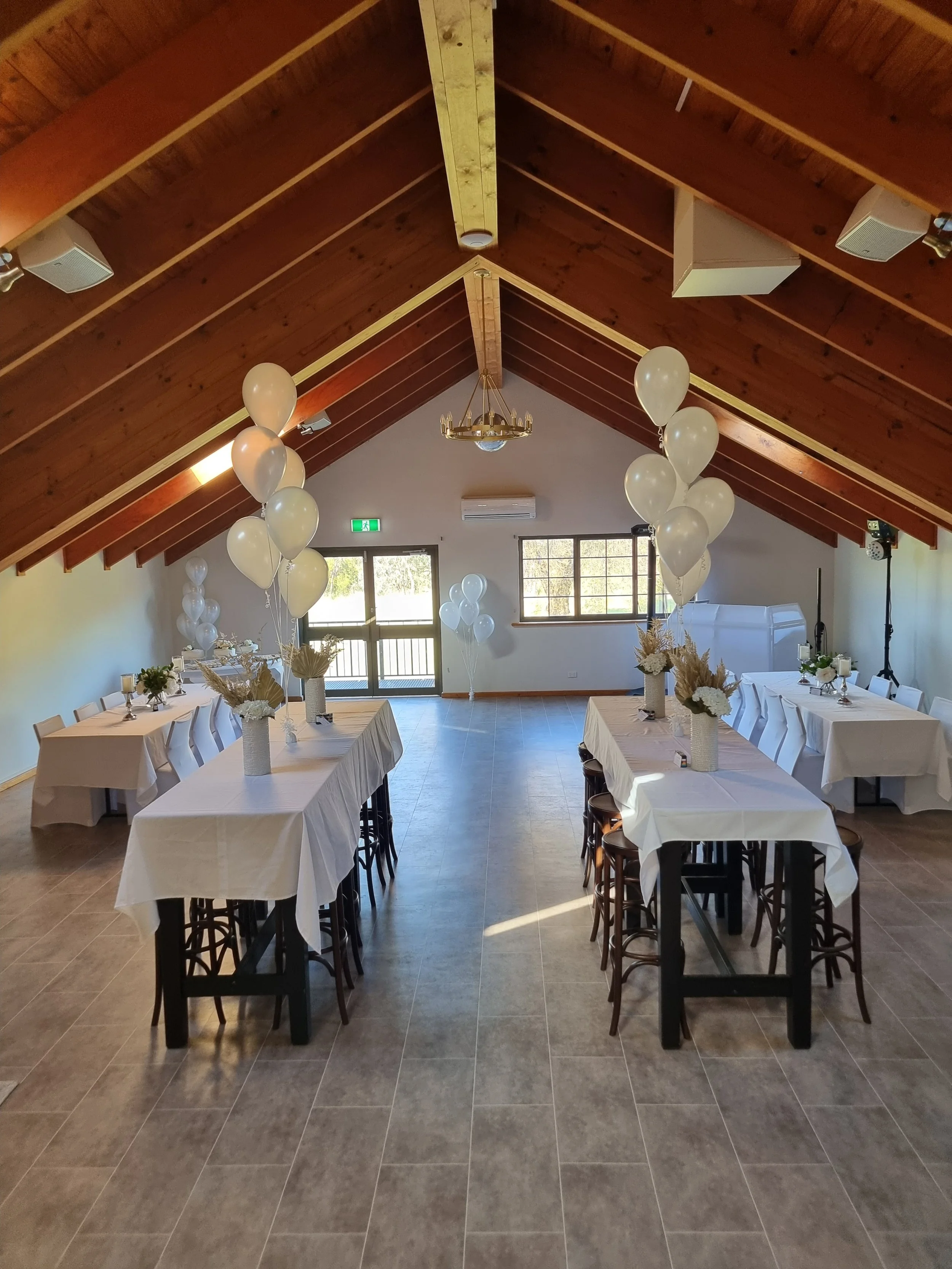 An elegantly decorated event hall with white tablecloths, white balloons, and floral centerpieces on the tables. The hall has a wooden vaulted ceiling, tiled floors, and large windows letting in natural light.