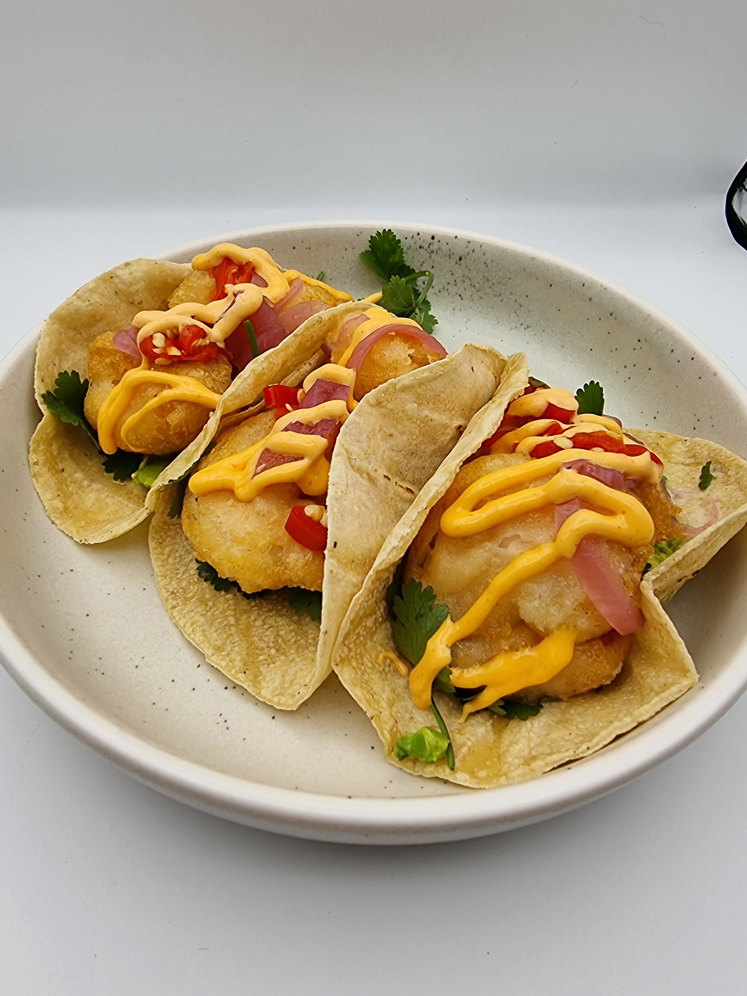Three fish tacos topped with chopped red chili, drizzled with yellow sauce, garnished with cilantro, served in soft corn tortillas on a white plate.