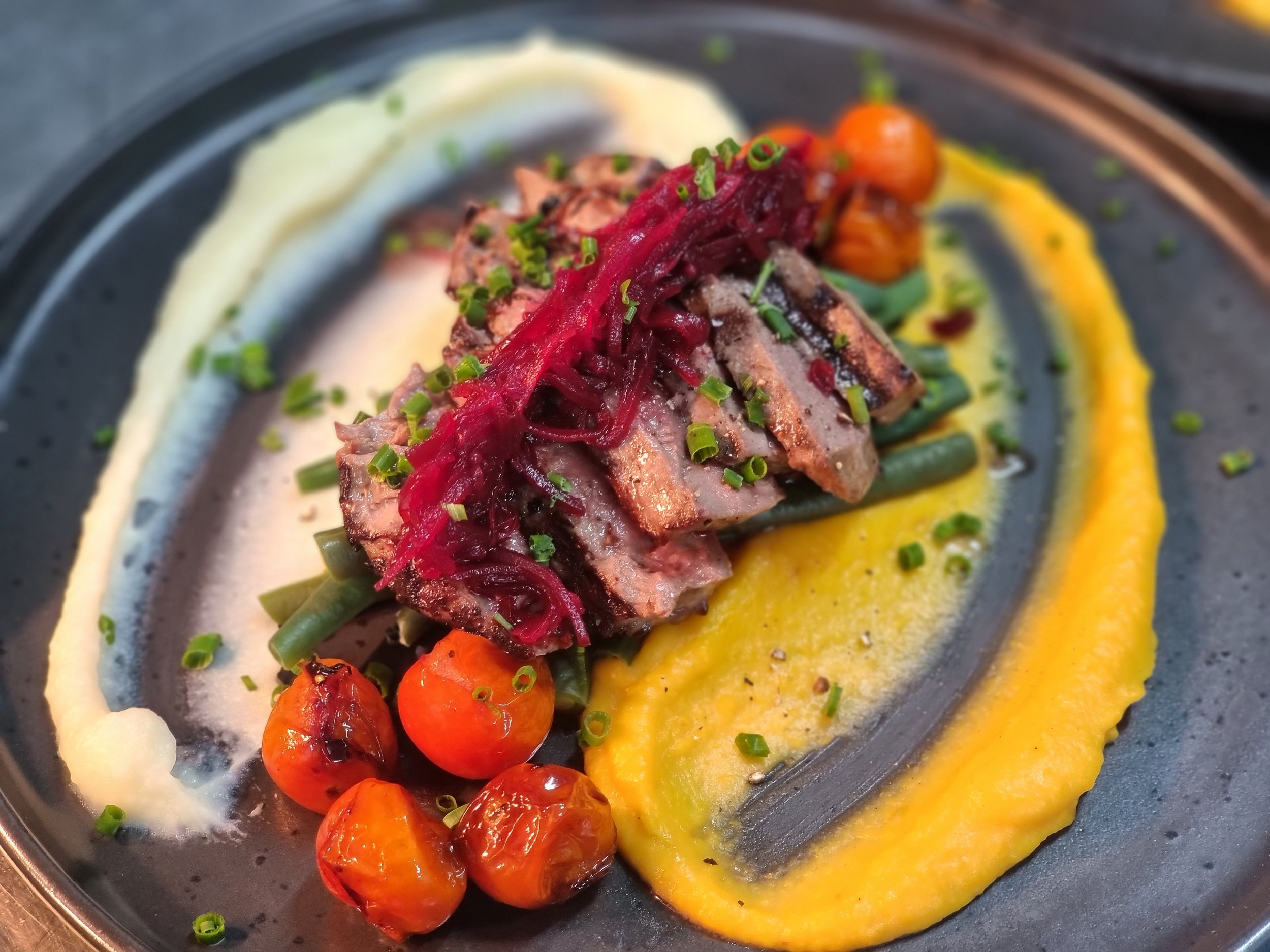 Artfully plated gourmet dish featuring slices of seared meat garnished with red pickled onions, served with roasted cherry tomatoes, green beans, and a colorful array of sauces including yellow, white, and black drizzles, on a dark plate.
