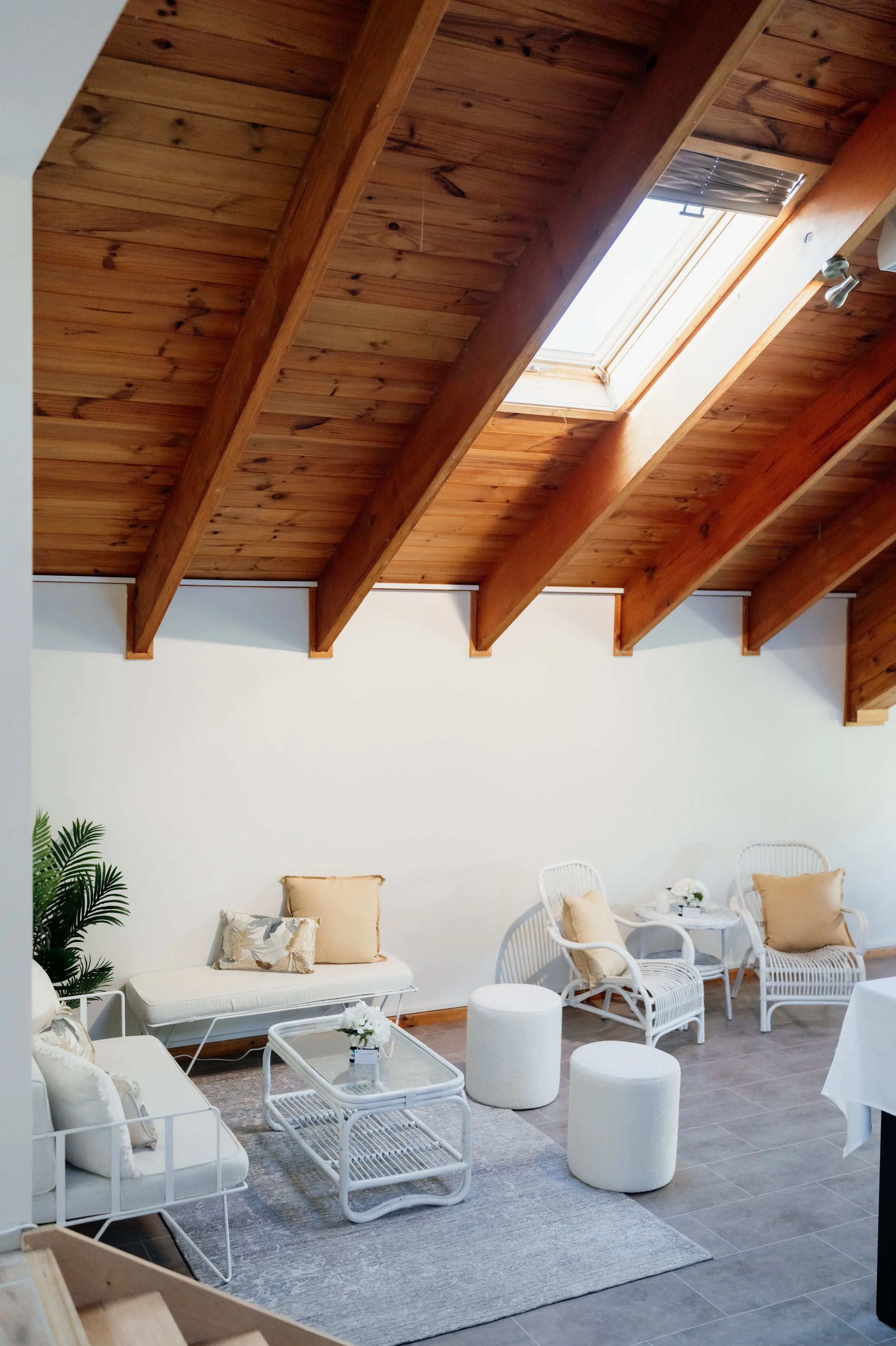 Living room with white furniture, beige pillows, wicker chairs, a glass coffee table, and a skylight in a room with wooden ceiling beams.