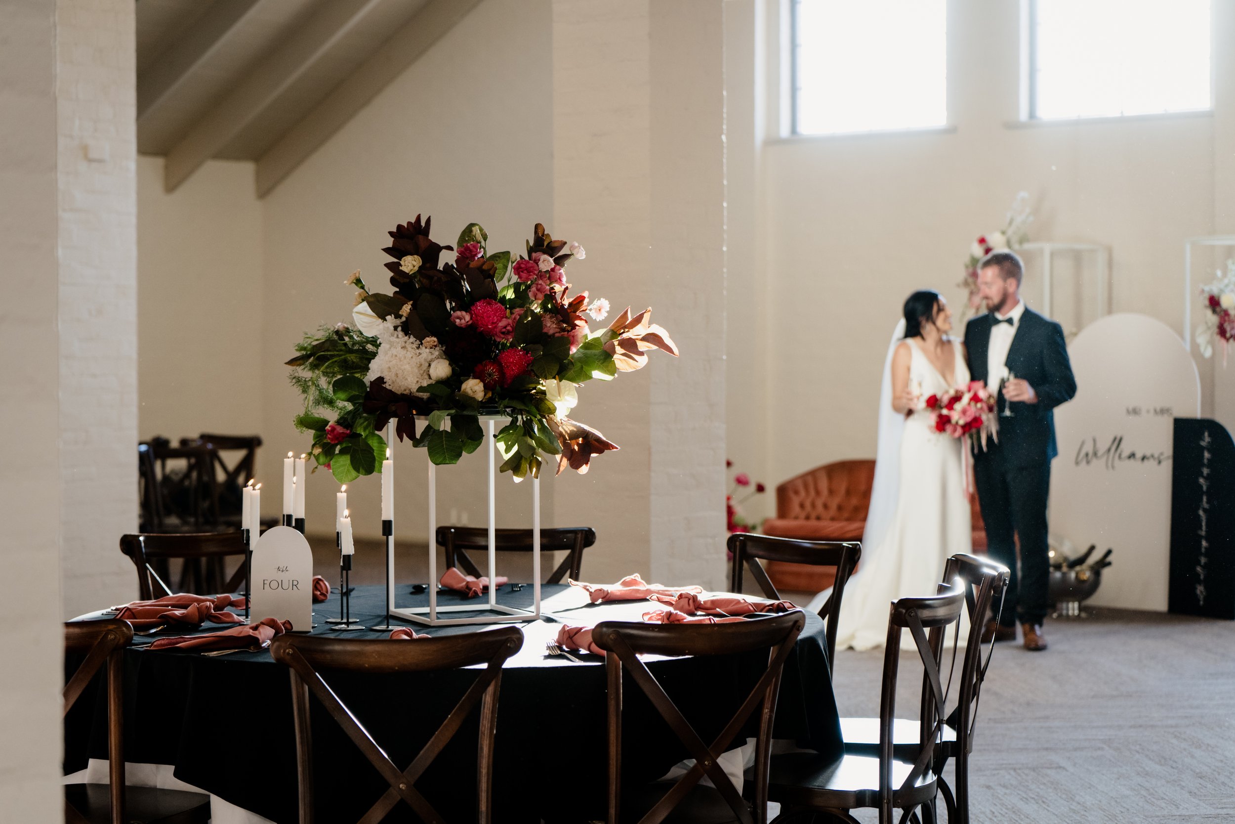 Wedding reception table with a floral centerpiece, pink napkins, and candles in a modern venue. In the background, a bride and groom are talking near the wall, with the bride holding a bouquet.