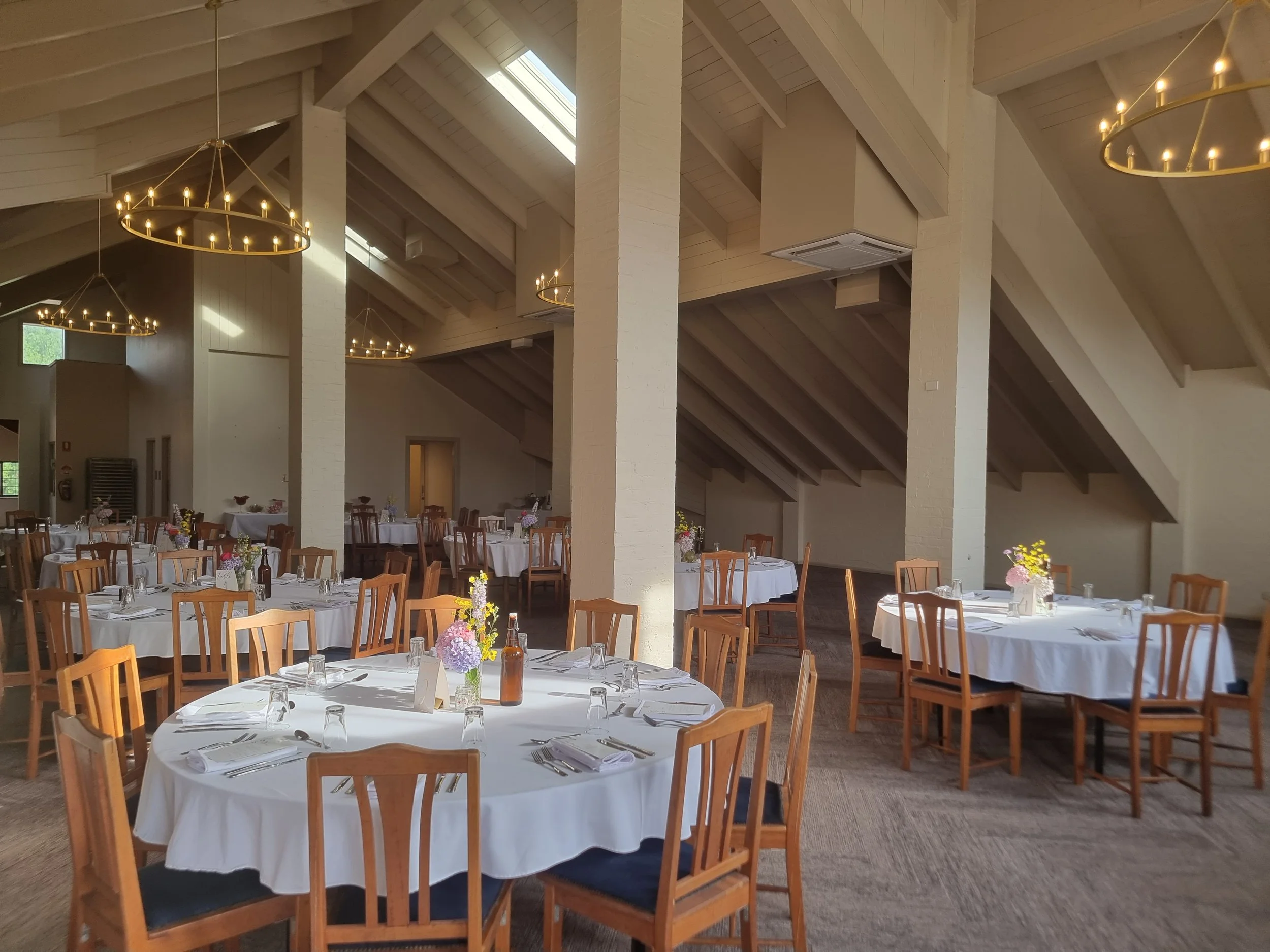 Elegant dining area with round tables covered in white tablecloths, set with glasses, silverware, and napkins. Each table has a floral centerpiece, and the room features wooden chairs, soaring ceilings with skylights, and modern circular chandeliers.