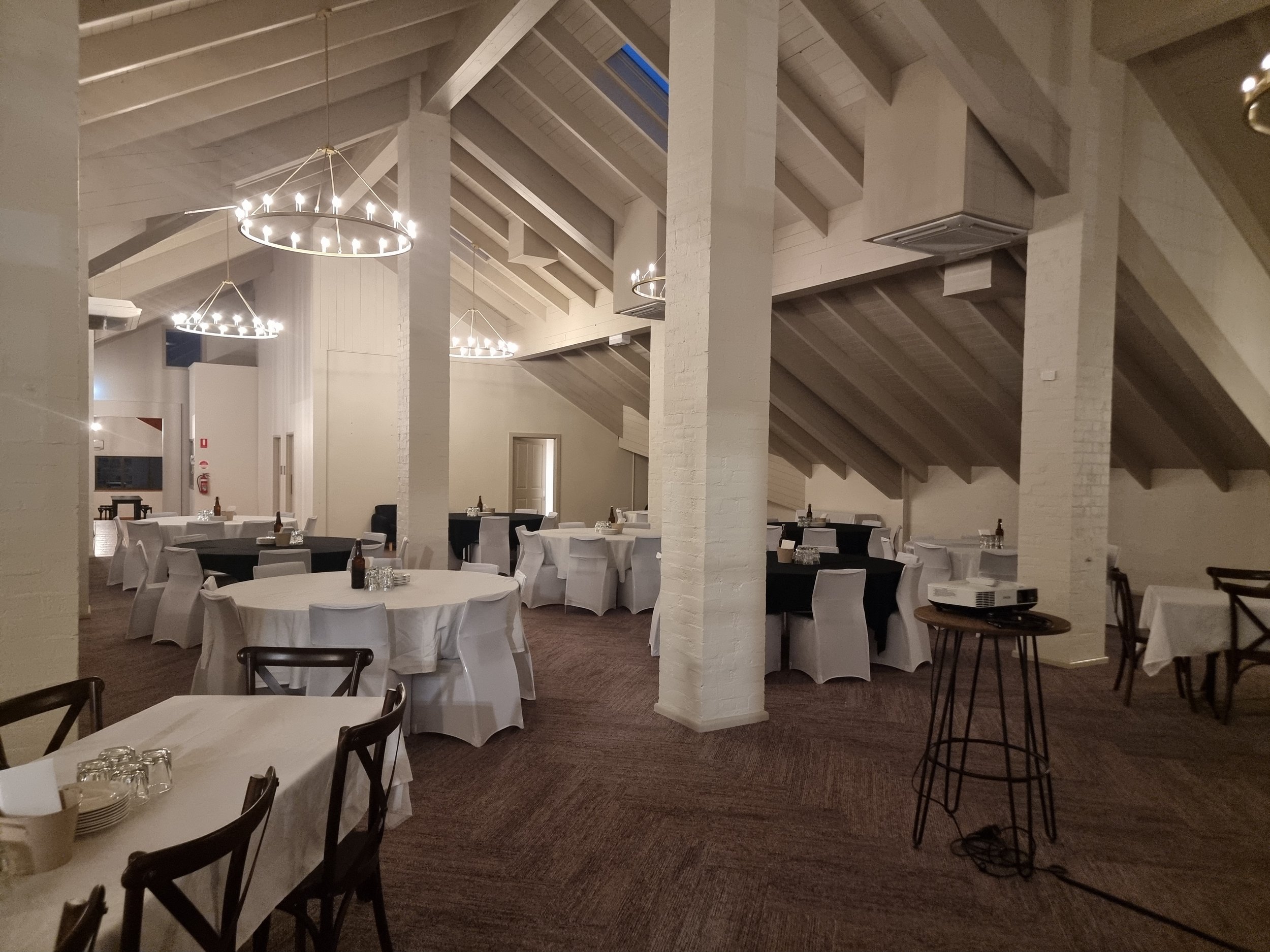 A spacious event hall with high, vaulted ceilings, white brick pillars, and elegant circular chandeliers. The room is set up for a formal gathering with round tables covered in white and black tablecloths, each with chairs, glassware, and bottles on