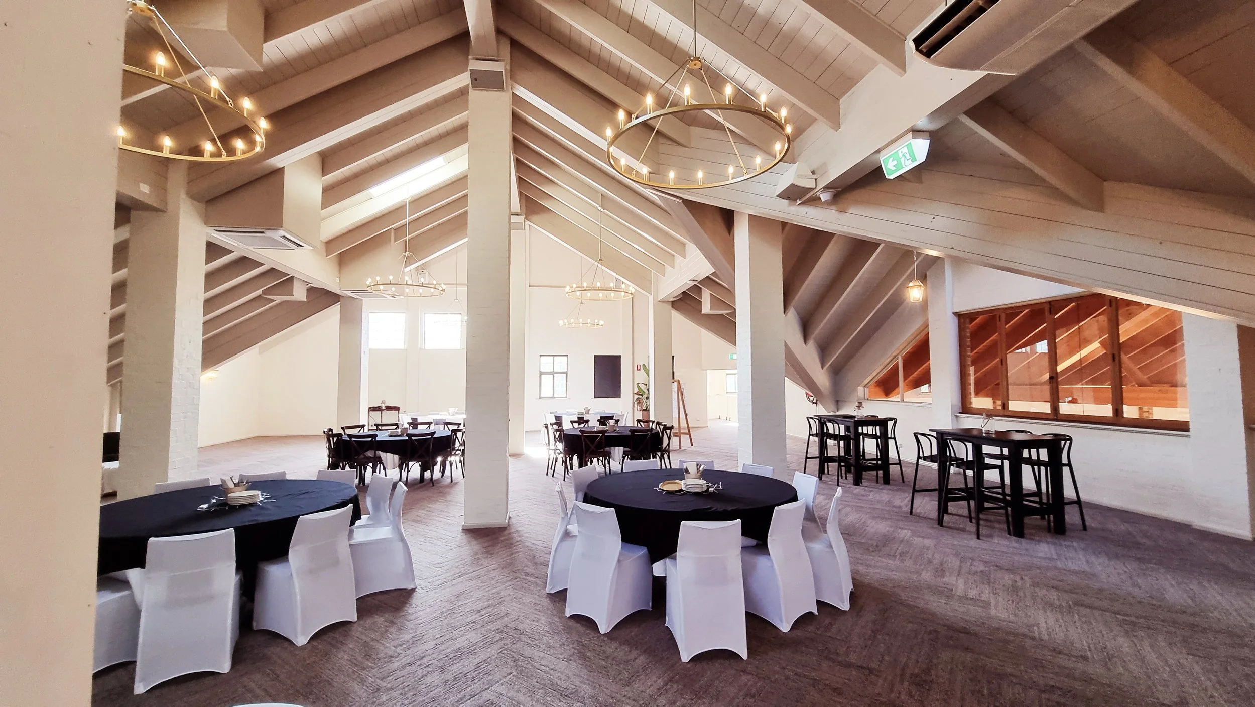 Event hall with round tables covered in black and white tablecloths, chairs with white slipcovers, high ceilings with wooden beams, chandeliers, and large windows.
