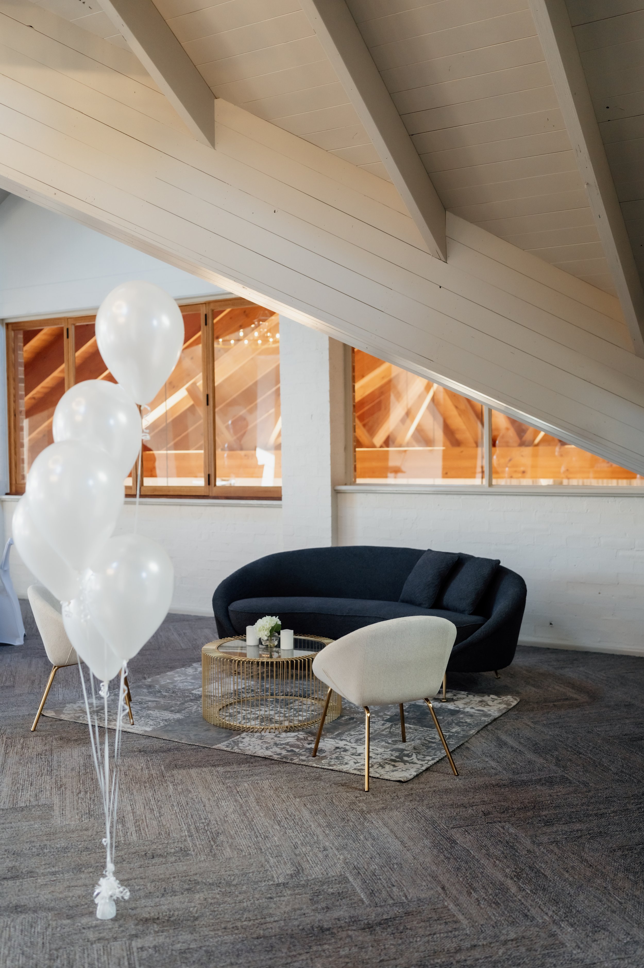Interior of a modern living room with a dark curved sofa, two light-colored chairs, a round gold coffee table, white candles, and a small vase with flowers, with white balloons, a wood-paneled wall, and a sloped ceiling.