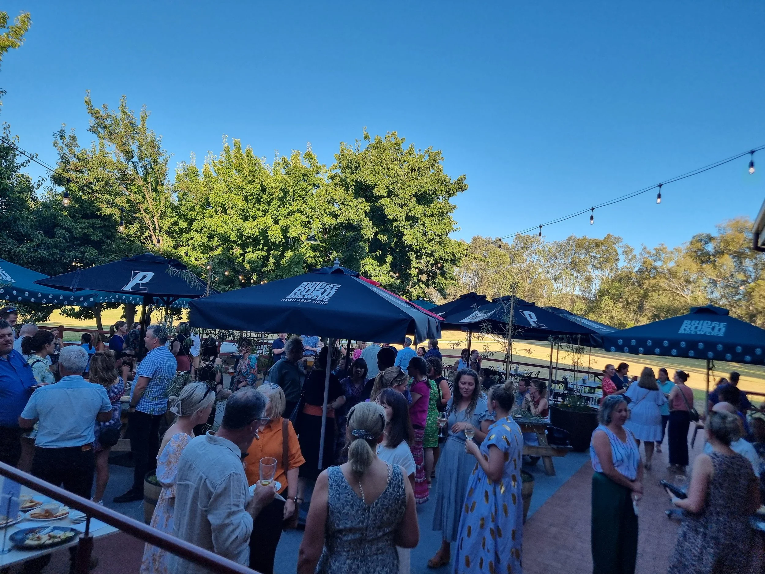 Crowd of people socializing outdoors at a party or event under large umbrellas with trees and string lights overhead.