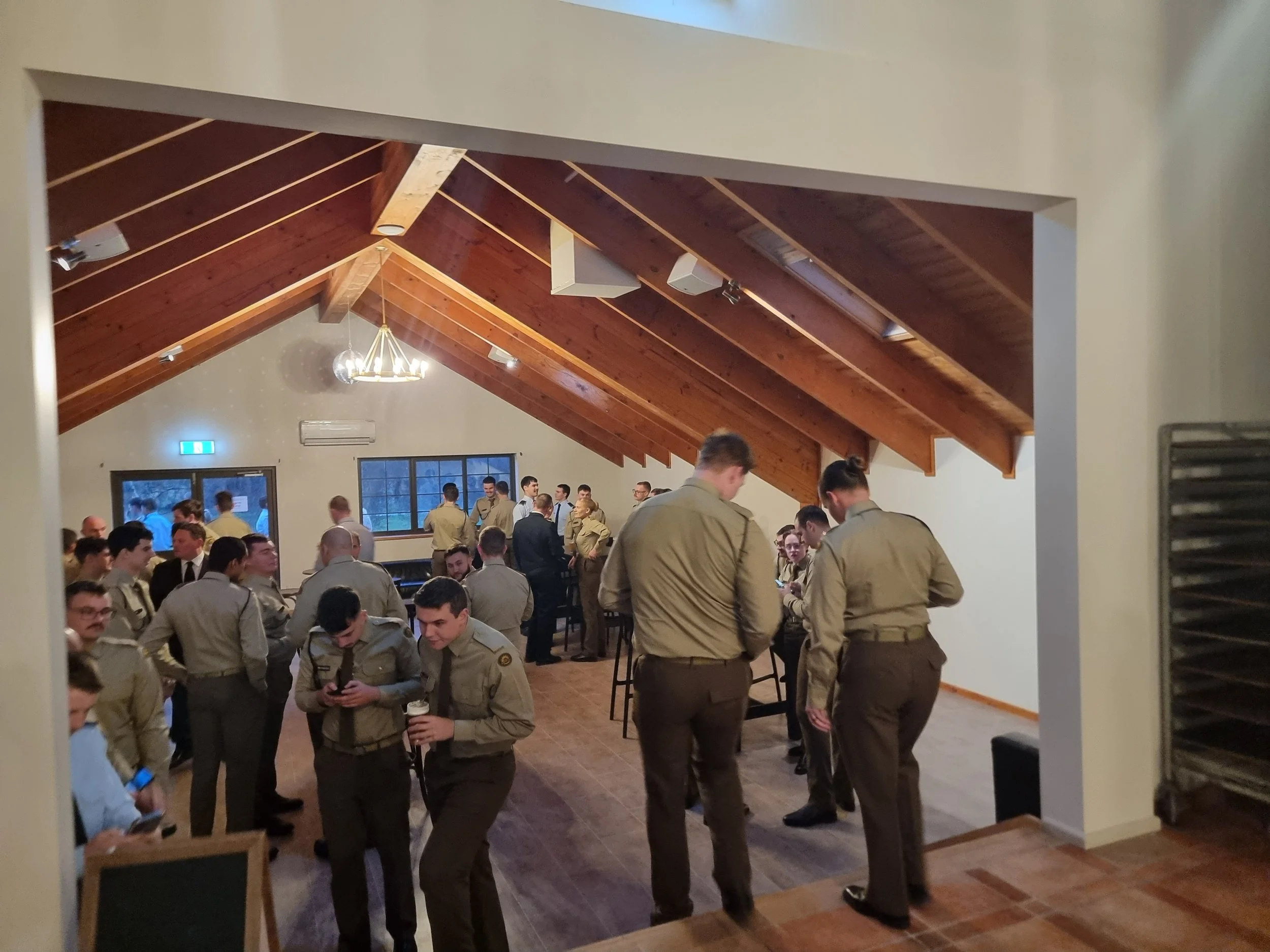Group of people dressed in military uniforms socializing in a room with wooden vaulted ceiling and windows.