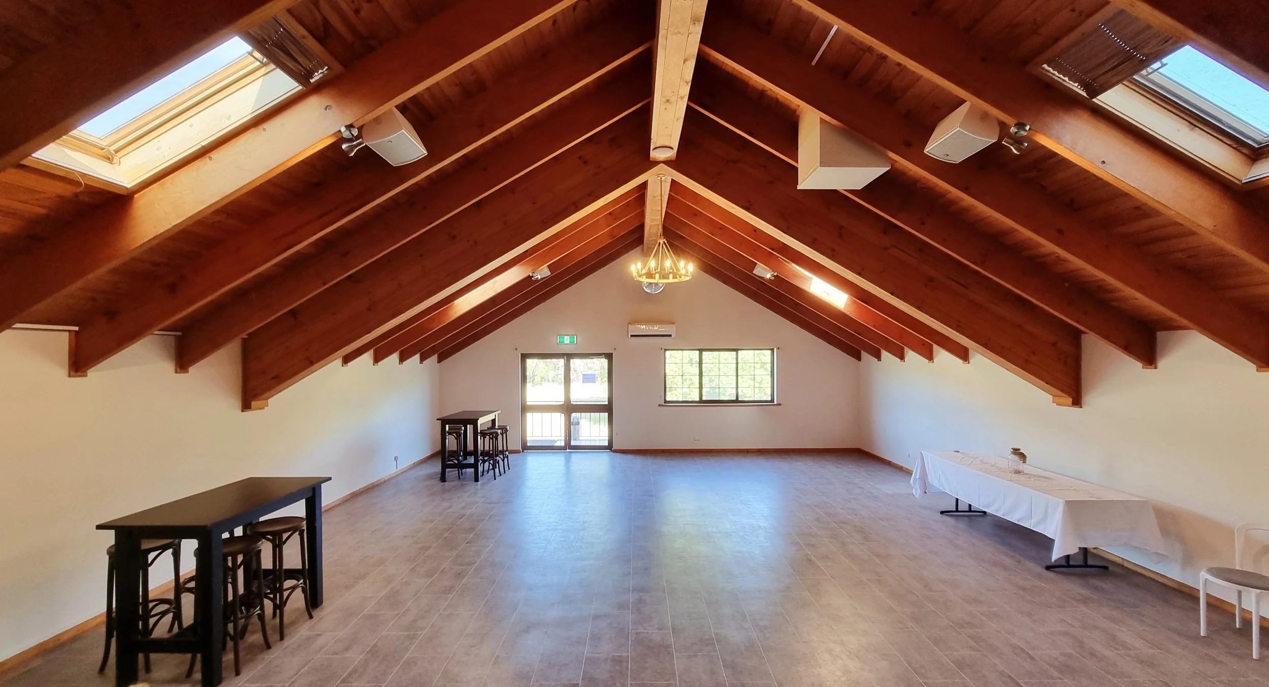 Empty event hall with wooden vaulted ceiling, skylights, chandelier, large window, glass door, and minimal furniture including tables and chairs.