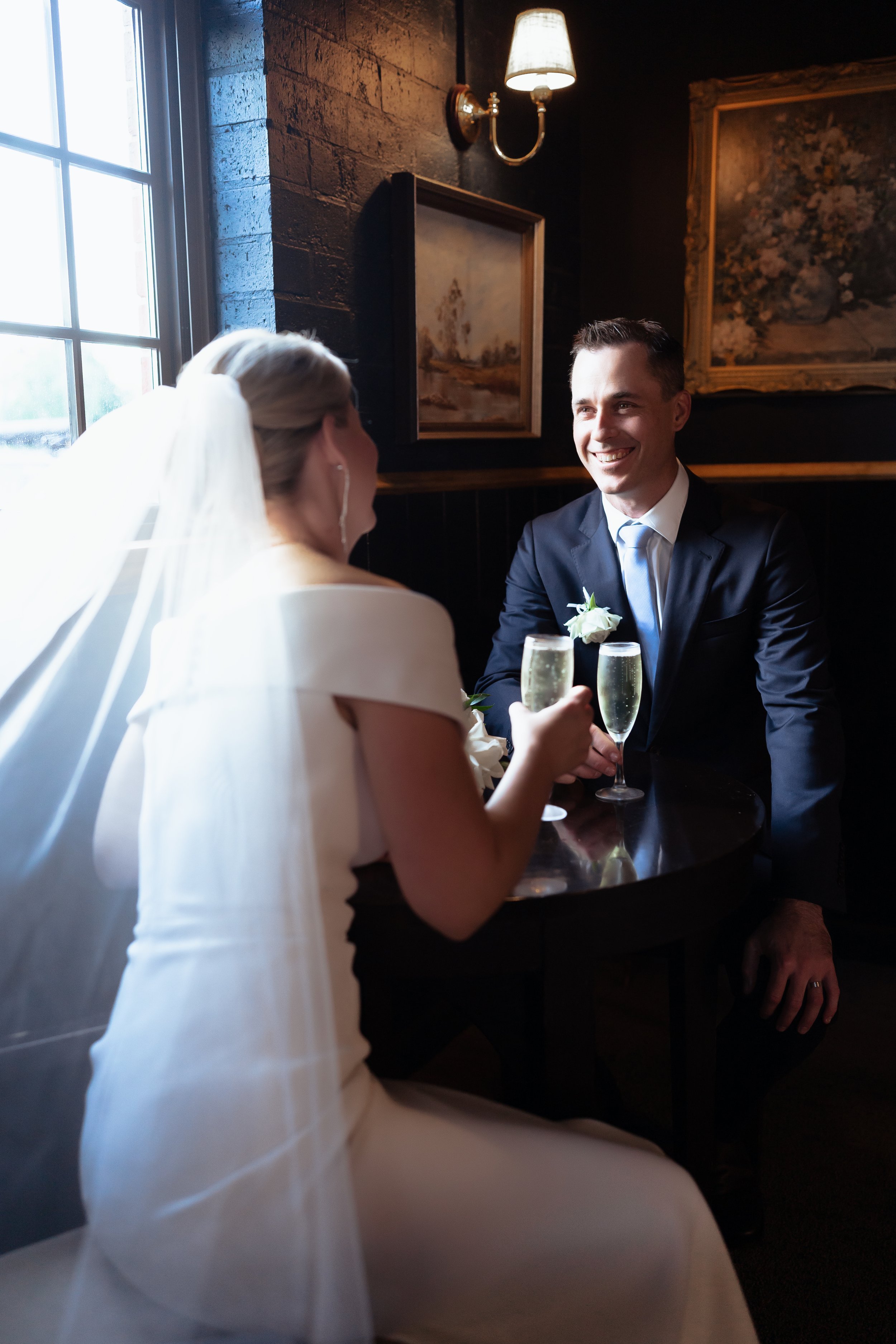 A bride and groom sitting at a small round table, holding champagne glasses, smiling at each other in a dimly lit room with dark walls, framed paintings, and a window letting in natural light.