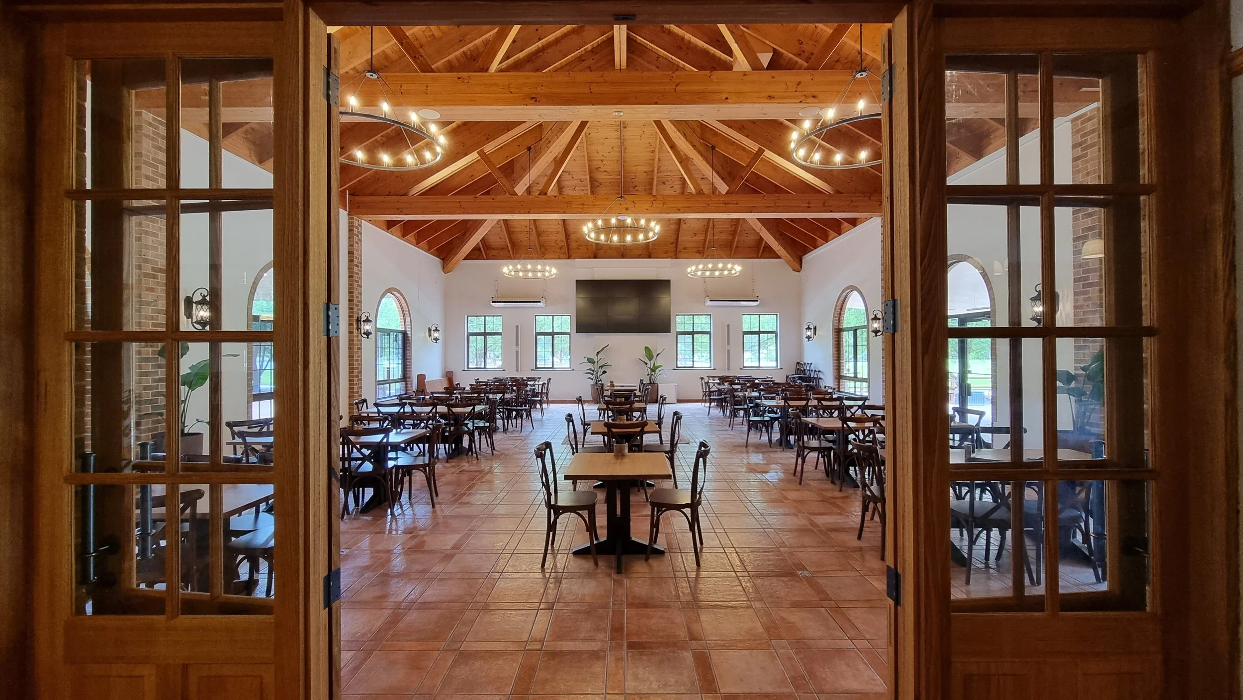 View of a spacious, well-lit dining area with wooden ceiling beams, multiple circular chandeliers, large windows, and arranged tables and chairs, seen through wooden-framed glass doors.