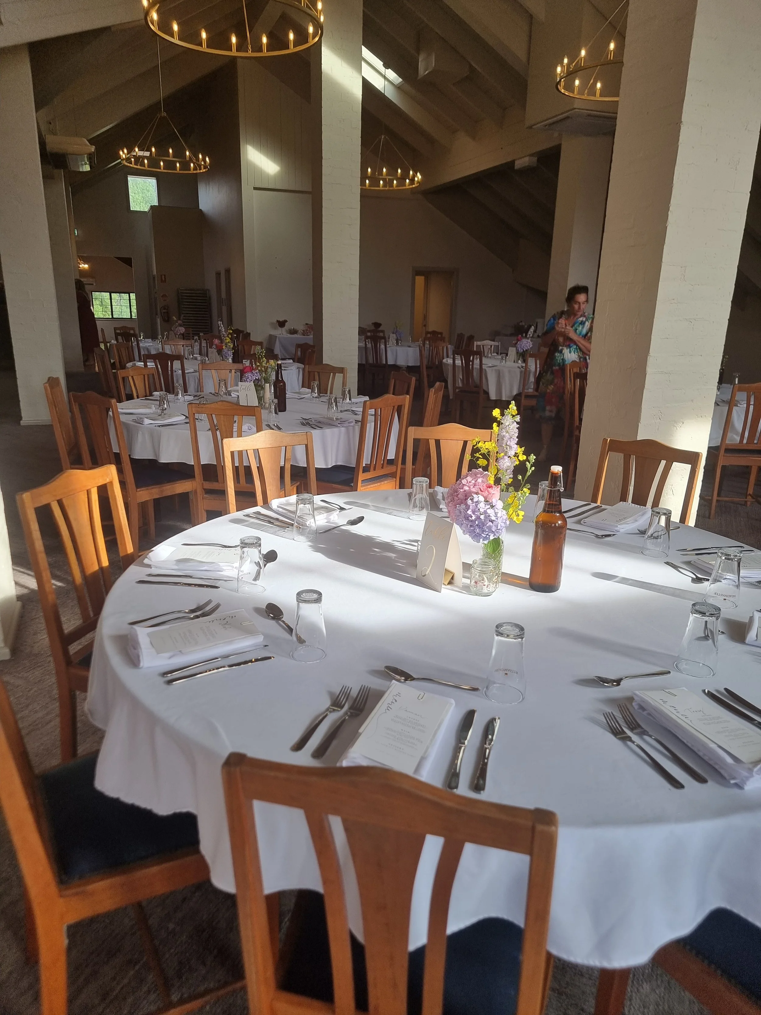 Round banquet table decorated with a white tablecloth, set with silverware, glasses, napkins, a menu, and a centerpiece with pink, purple, and yellow flowers, in a spacious dining hall with multiple similar tables, wooden chairs, and chandeliers.