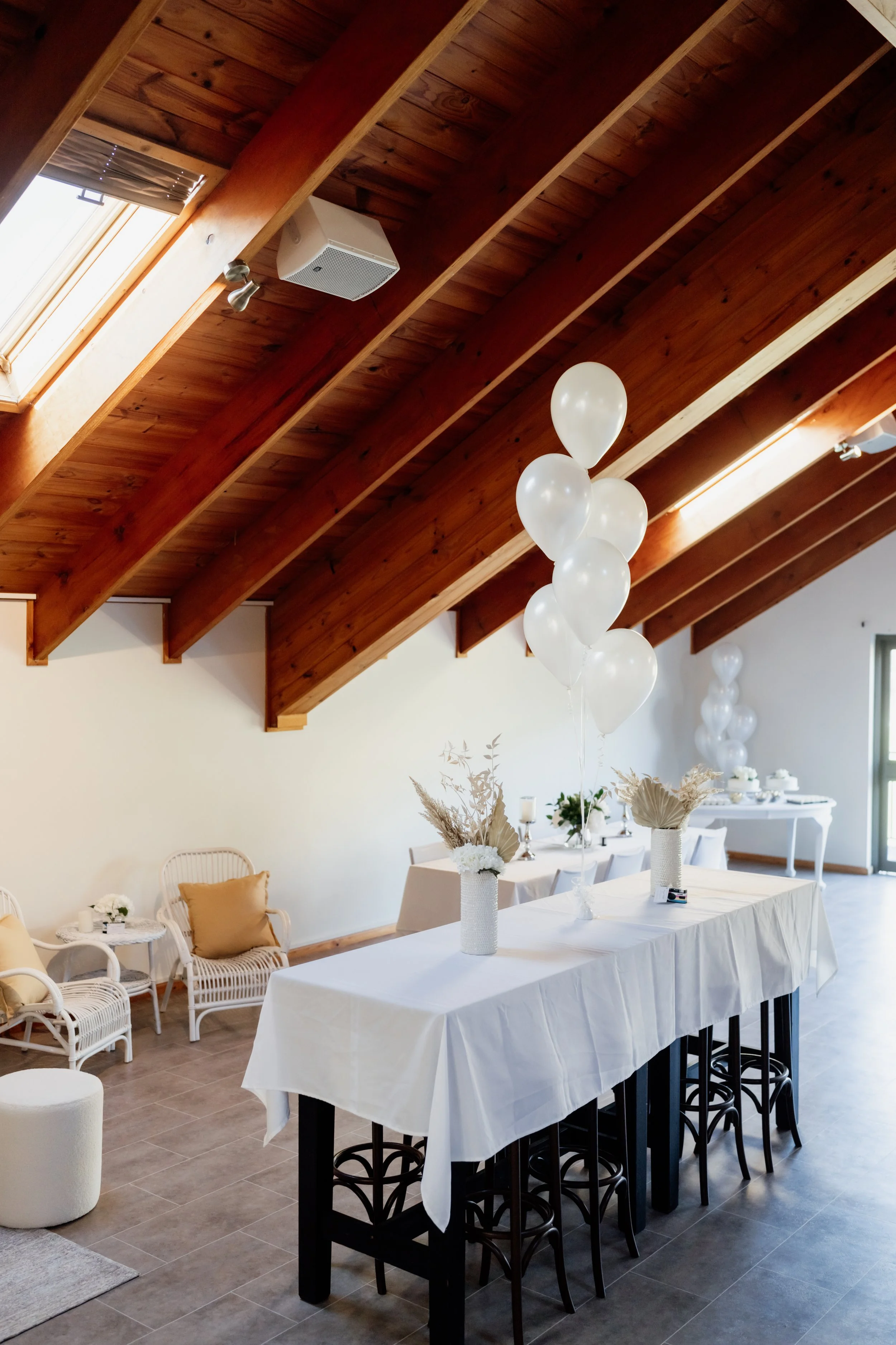 Decorated indoor event space with white balloons, wicker chairs, white table with vases of dried flowers, and a long table with additional balloons in the background. Wooden ceiling with skylights.