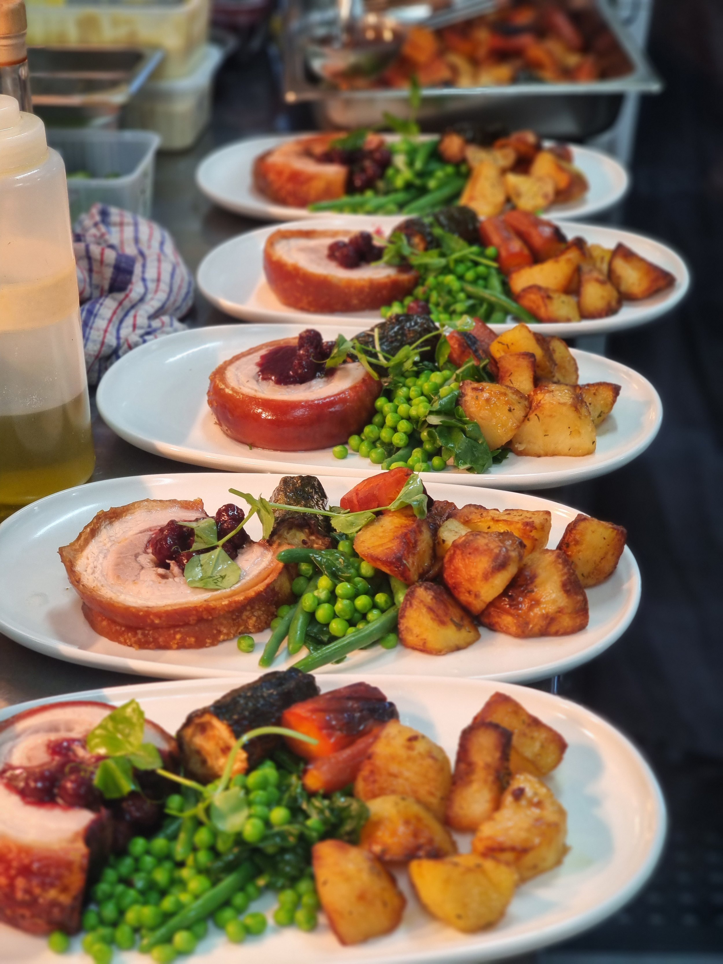 Multiple plates of rolled pork with sauce, roasted potatoes, green peas, and green beans, lined up on a table.