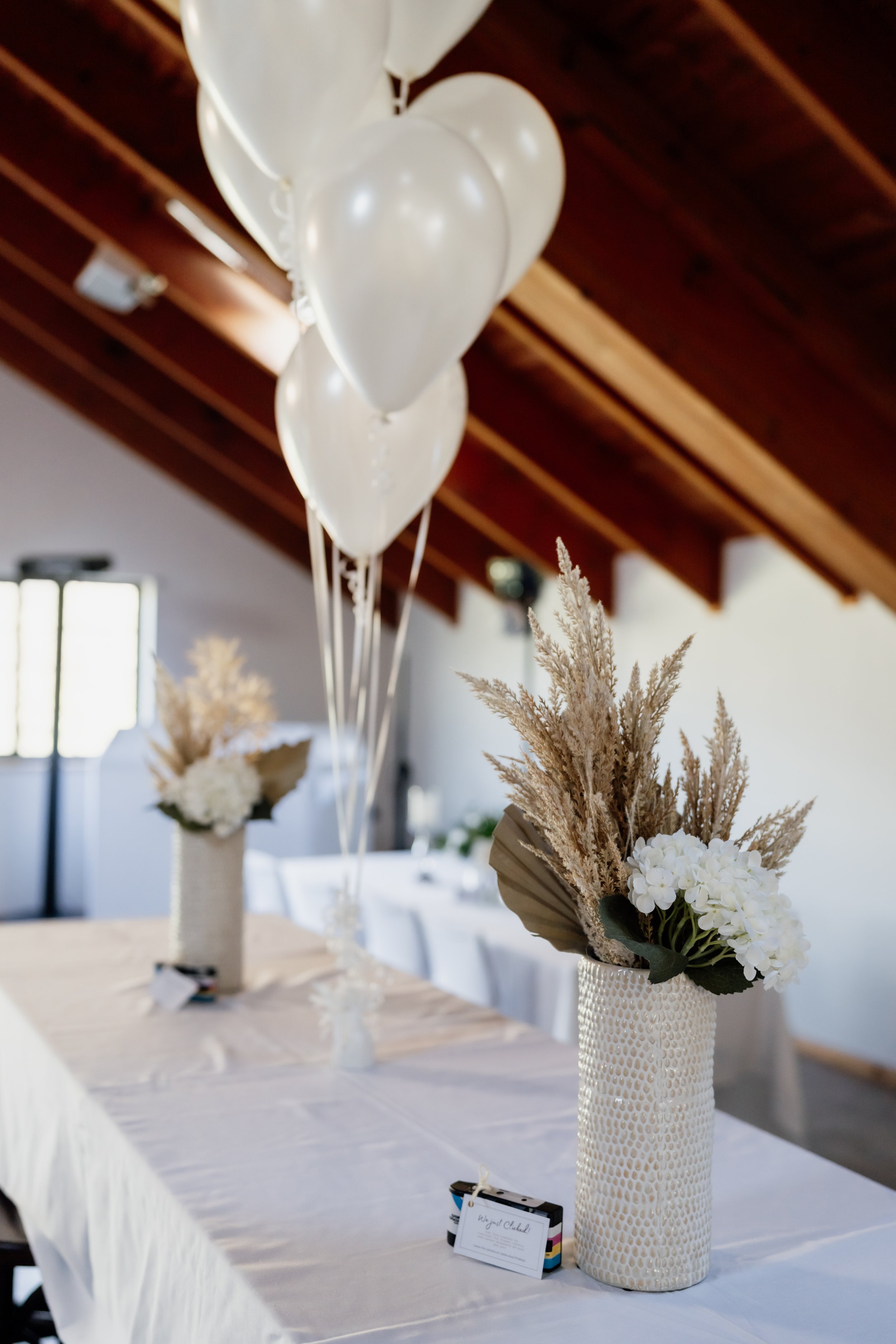 Table decorated with white floral arrangements in textured vases, white balloons, and place cards in a well-lit room with a wooden ceiling.