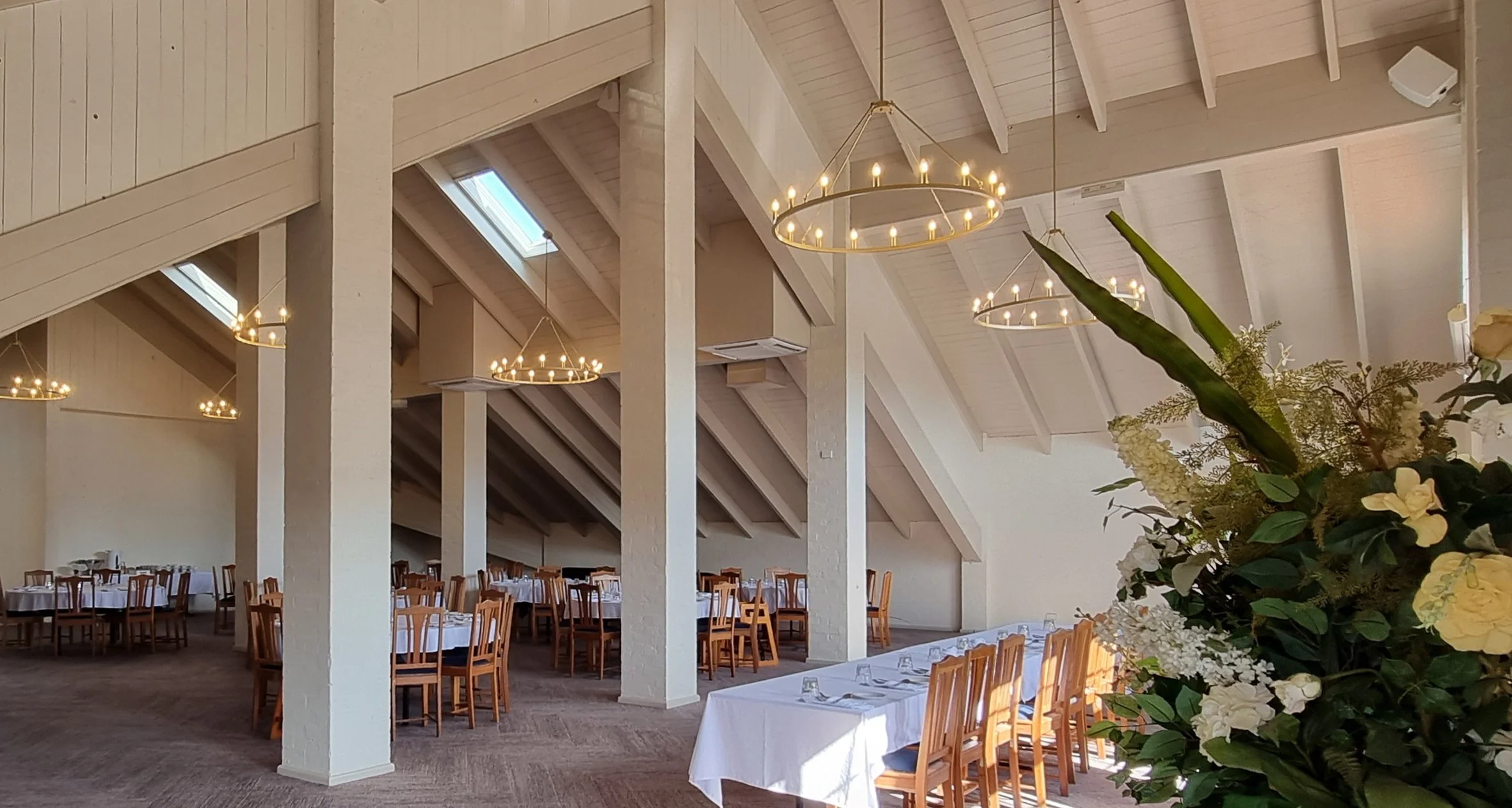 Elegant banquet hall decorated with white tablecloths, wooden chairs, floral arrangements, and circular chandeliers, ready for an event.