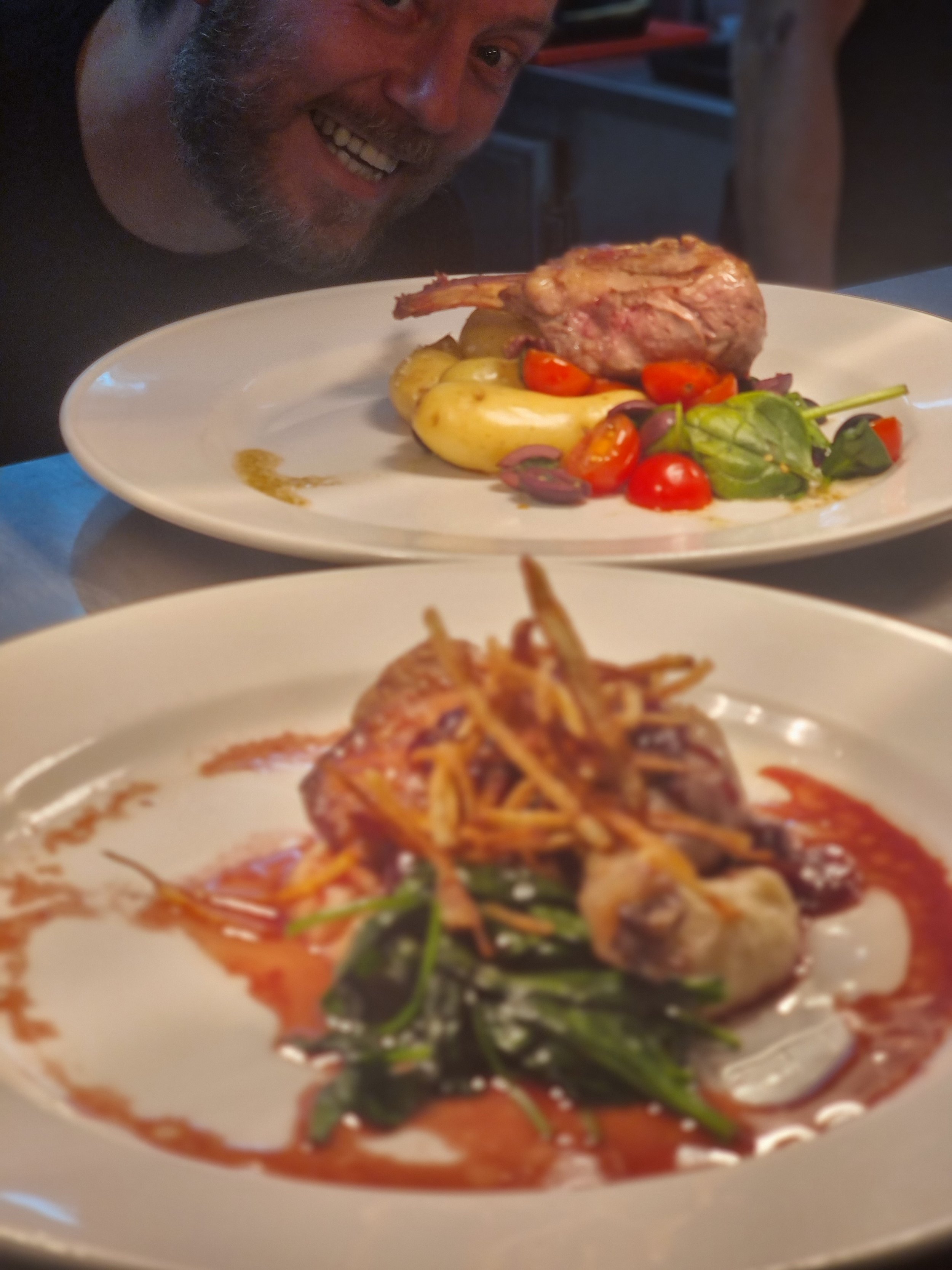 A man smiling at two plates of gourmet dishes, one featuring a cut of cooked meat with vegetables and greens, and the other with a plated meat dish with sauce and garnishes.