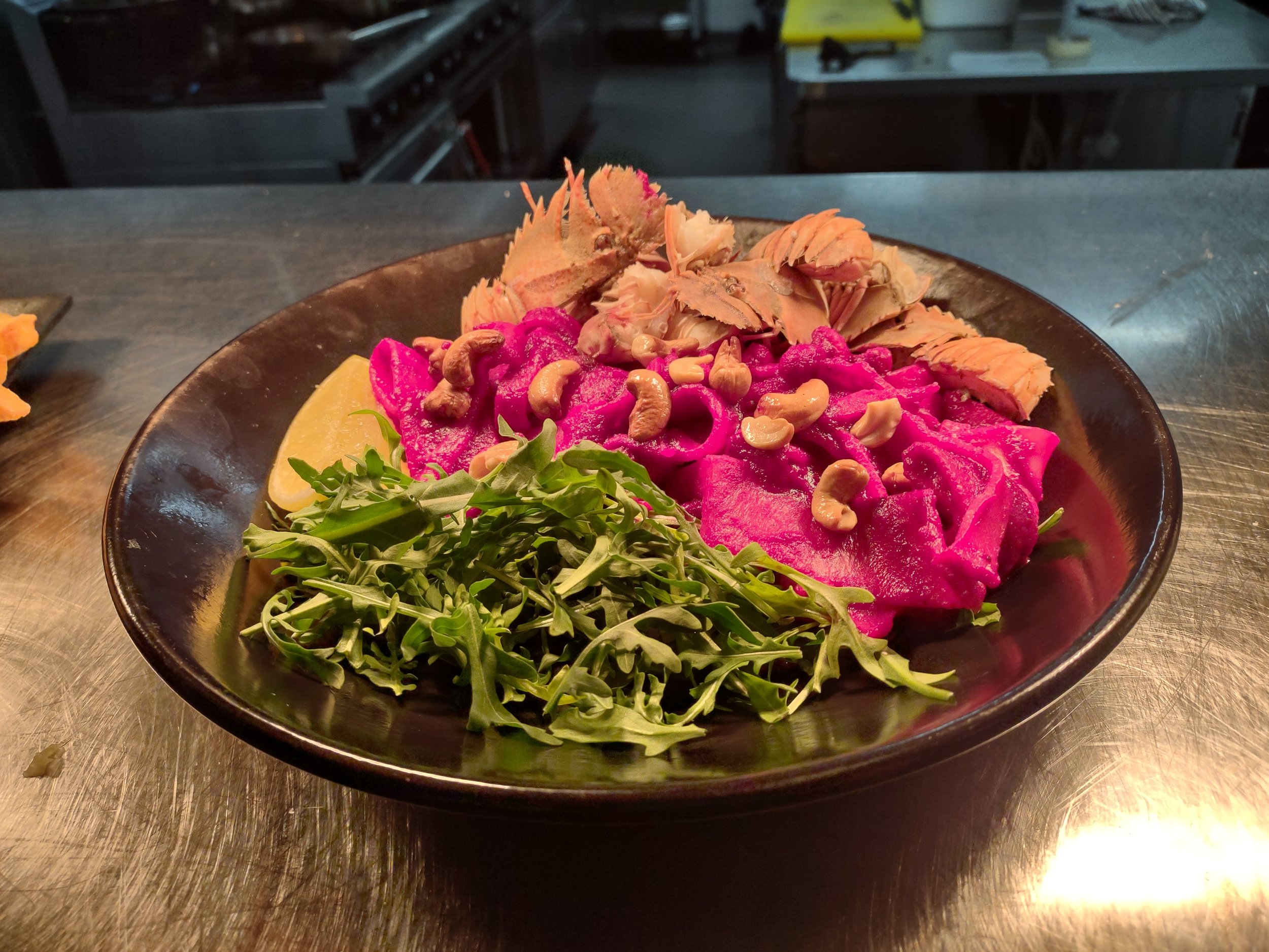 A black bowl containing bright pink beet hummus topped with cashew nuts, along with a lemon wedge, fresh arugula, and slices of salmon, on a stainless steel countertop in a kitchen.