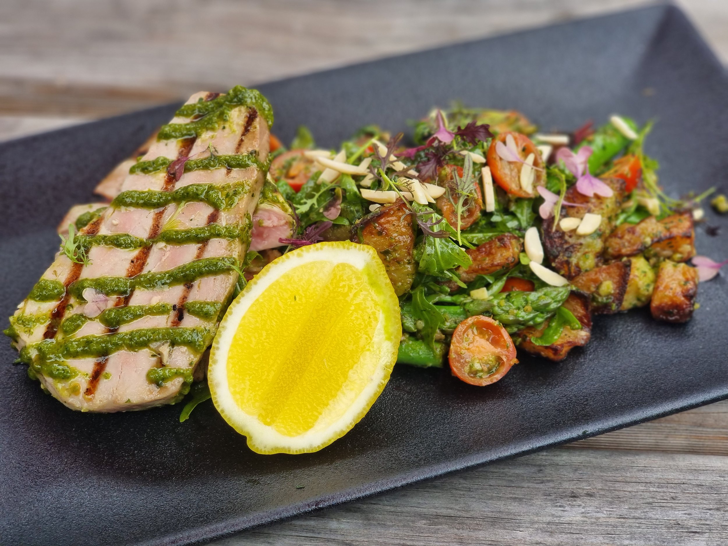 Grilled tuna steak with green sauce, side salad with cherry tomatoes, herbs, nuts, and a lemon wedge on a black rectangular plate.