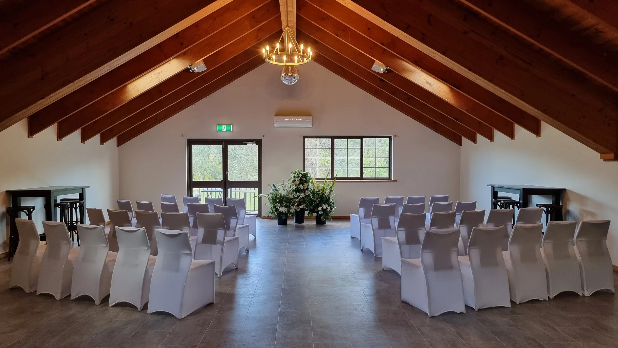 Empty wedding ceremony hall with white chairs arranged in rows, a floral arrangement at the front, wooden ceiling, large window, and glass door.