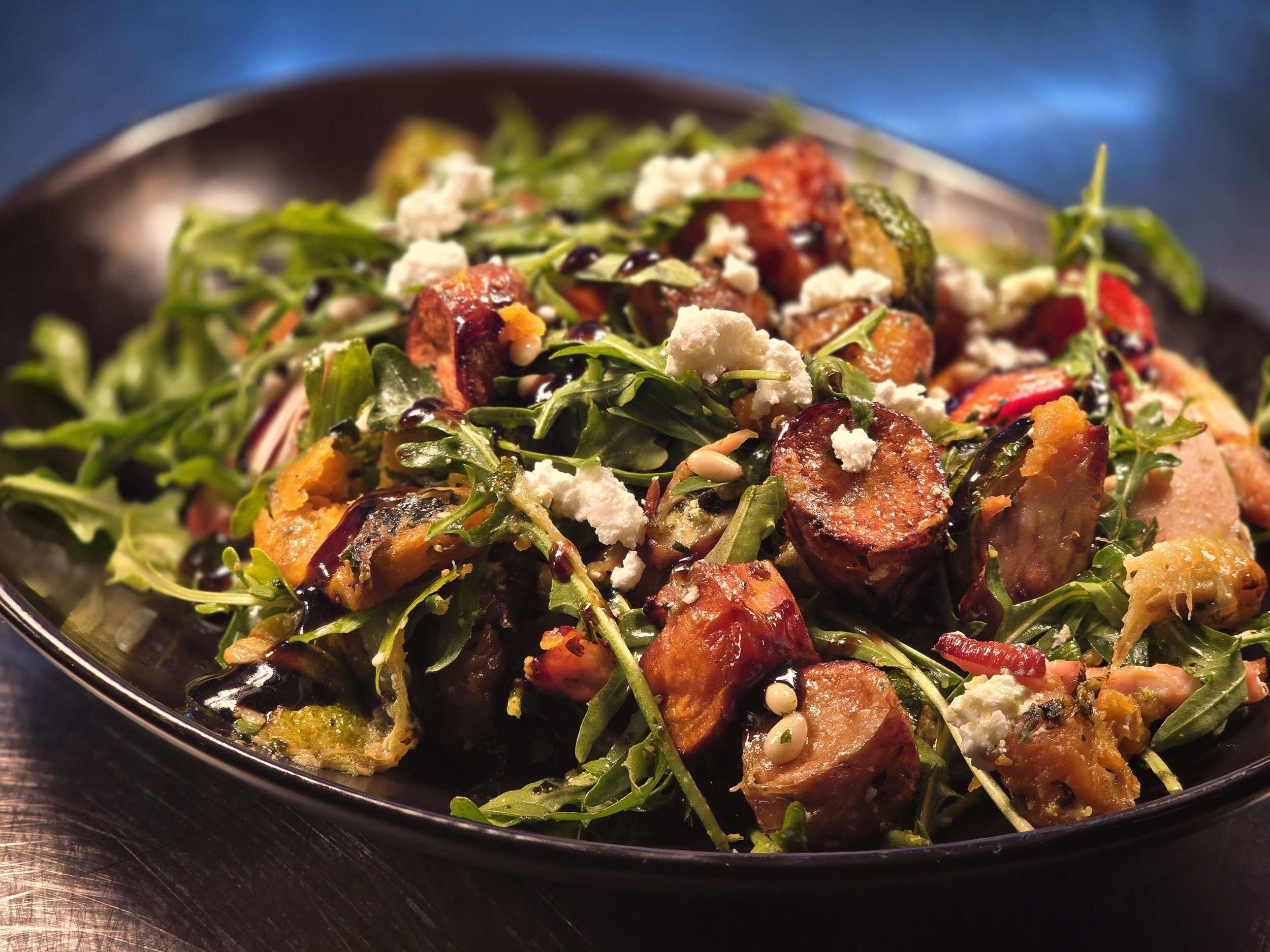 Close-up of a salad with roasted vegetables, arugula, crumbled cheese, and a drizzle of balsamic glaze in a black bowl