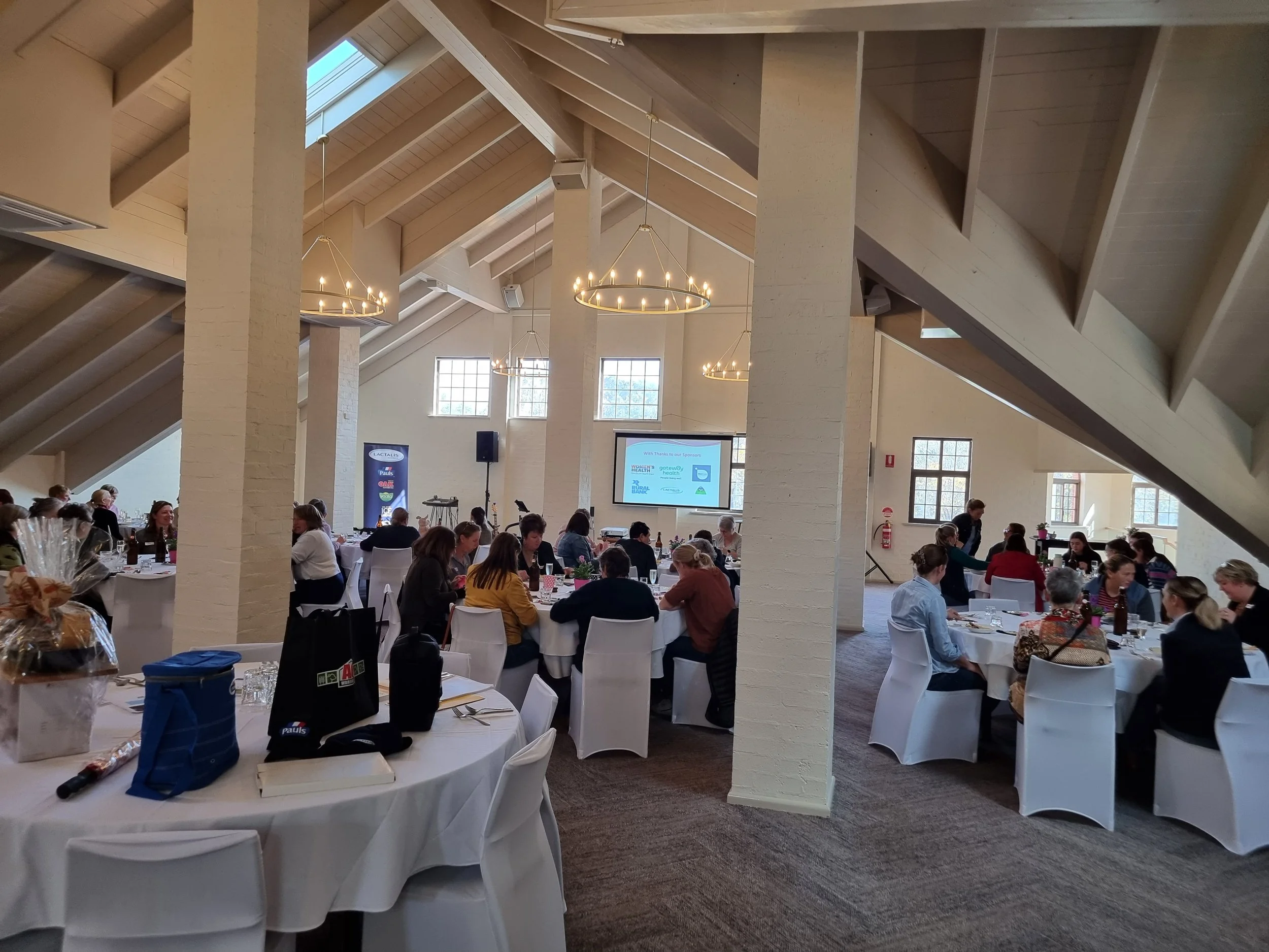 Conference room with round tables covered in white tablecloths, filled with seated attendees and event materials, under a high, vaulted ceiling with large windows. A presentation screen is visible at the front.