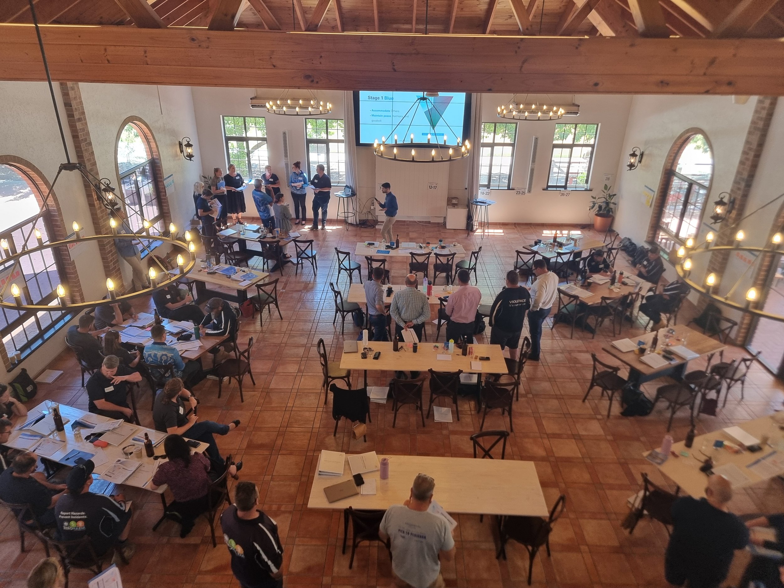 Interior of a spacious conference room with people gathered, some participating in a presentation and others seated at tables, sunlight streaming through large windows.