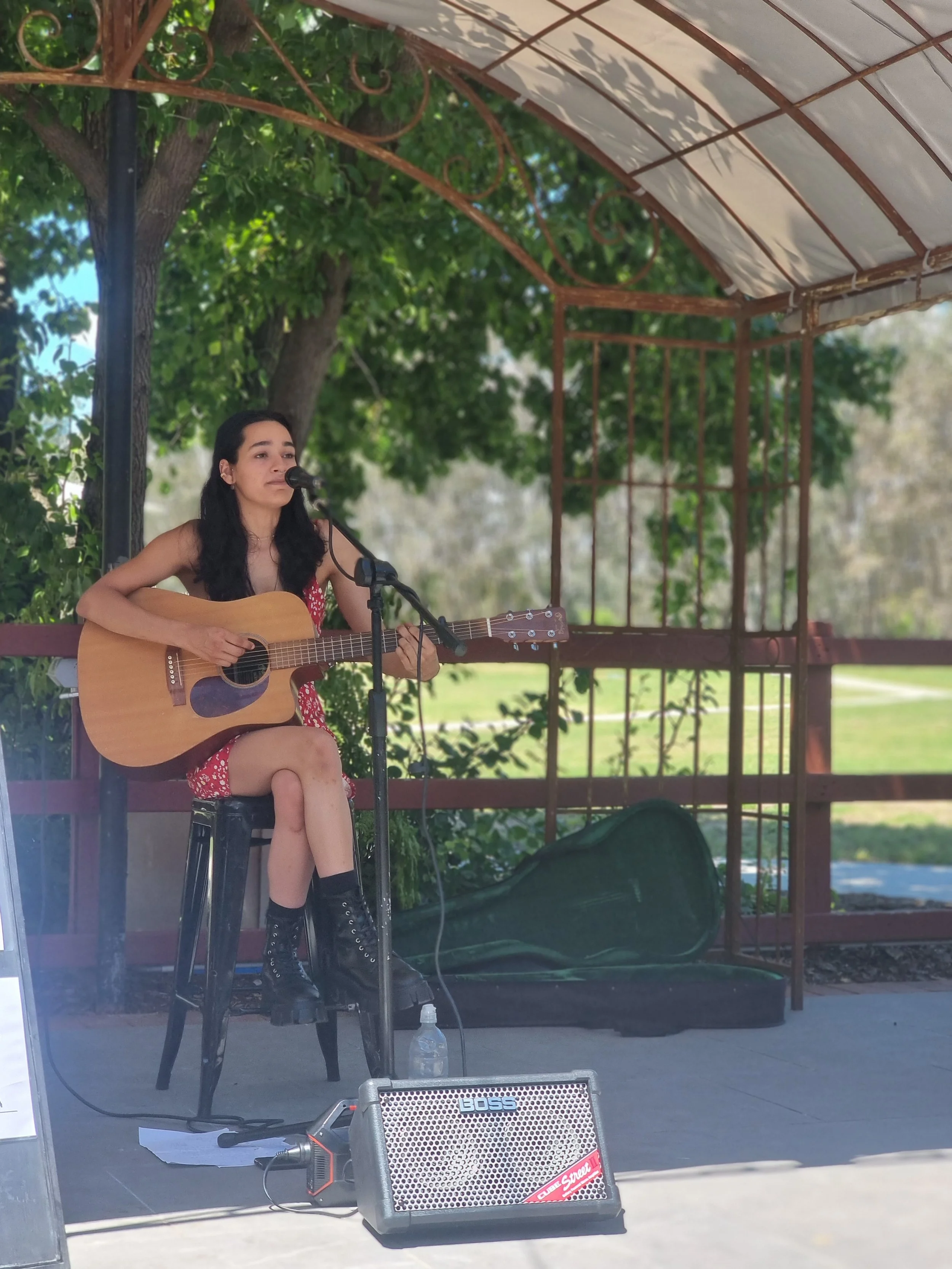 A young woman with long dark hair playing an acoustic guitar and singing into a microphone outdoors, seated on a tall black stool.