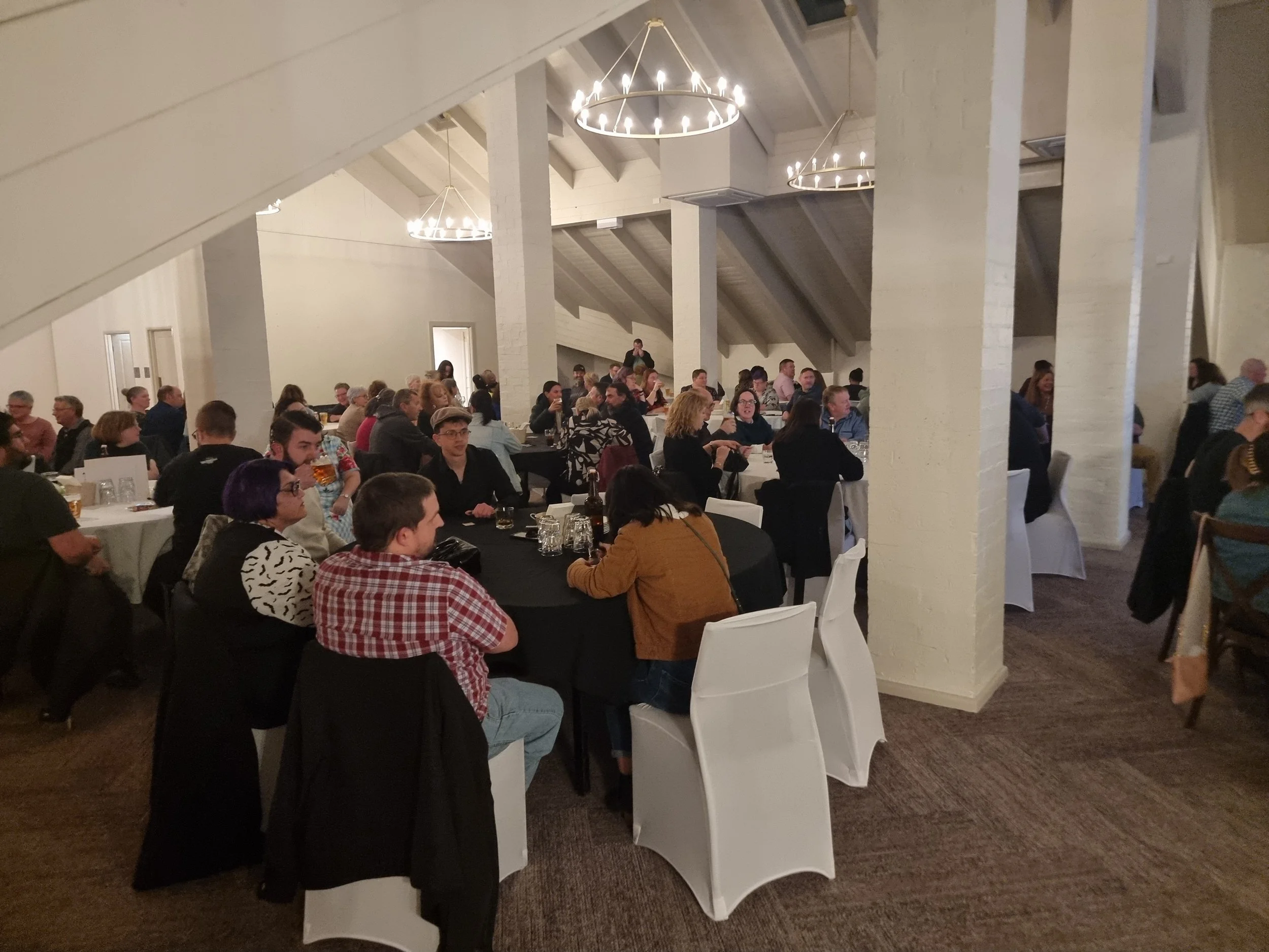People seated at round tables with black and white tablecloths in a spacious, white-painted event hall with high vaulted ceilings and modern chandelier lighting.