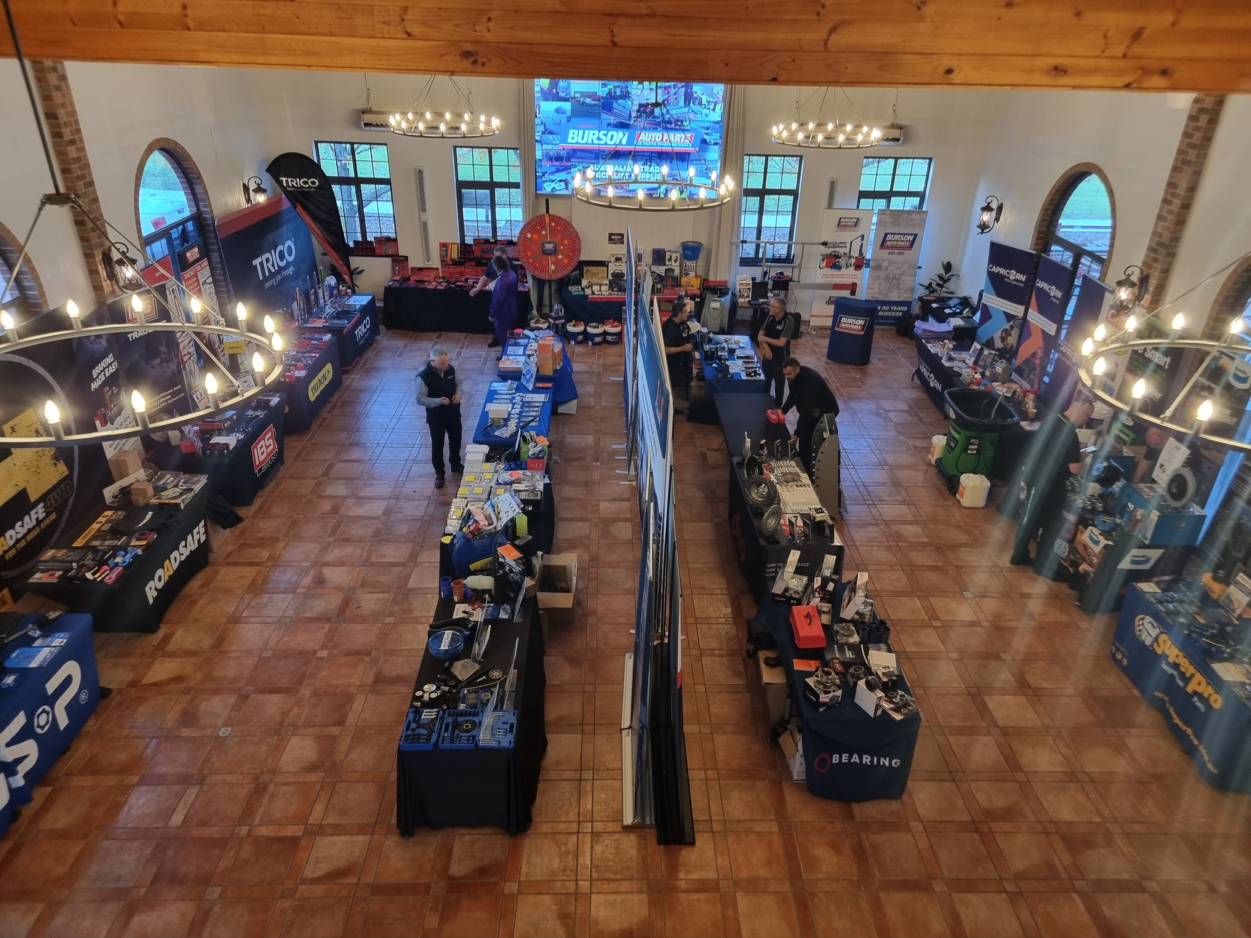 Overview of an indoor trade show with various vendor booths and tables displaying products, including automotive parts and tools, under warm lighting. People are walking and browsing.