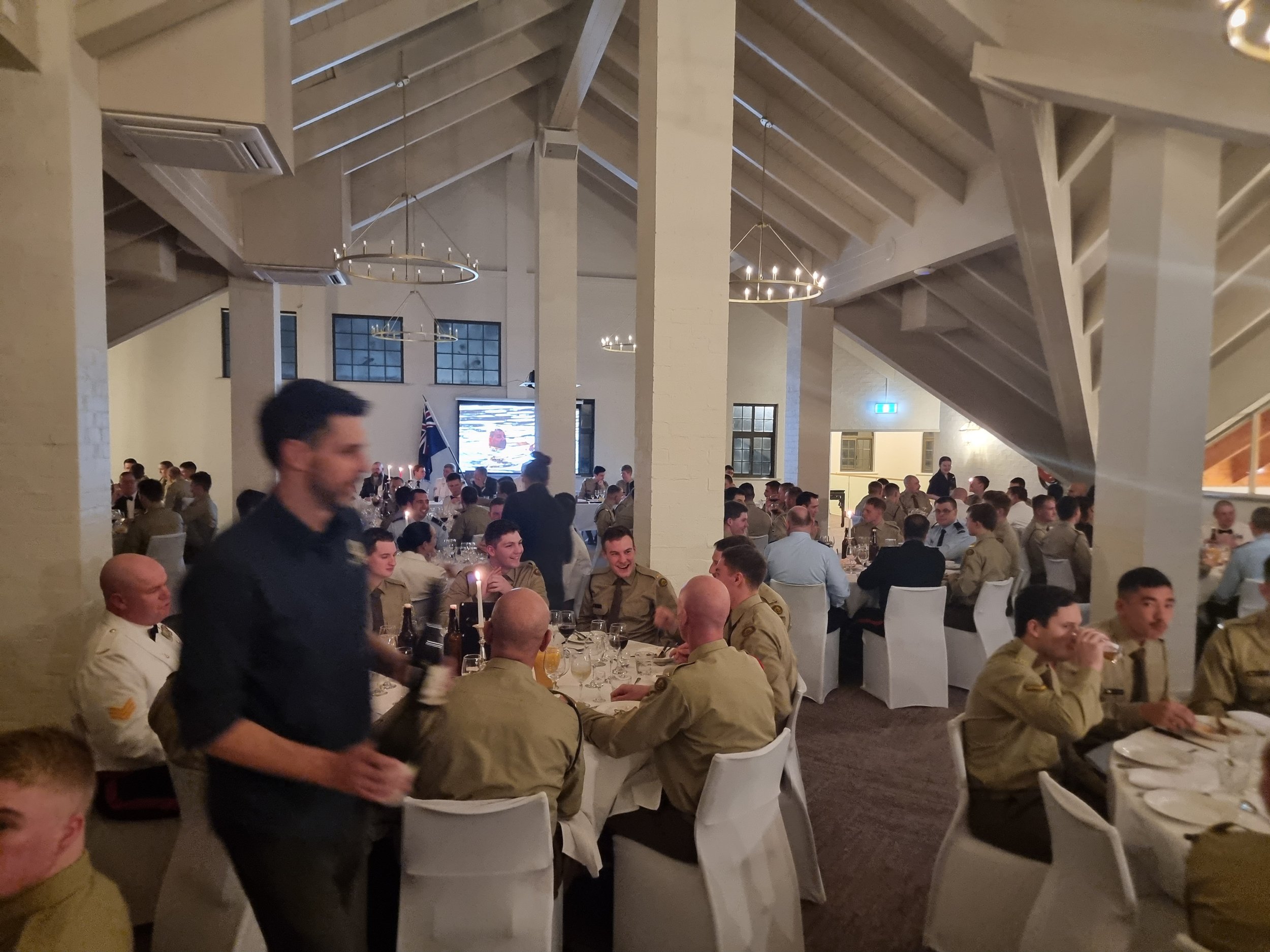 A large group of military personnel in uniform seated at round banquet tables in a spacious, well-lit dining hall with white walls, high sloped ceilings, and modern chandeliers. A waiter in a black shirt is walking past, holding bottles, and there is