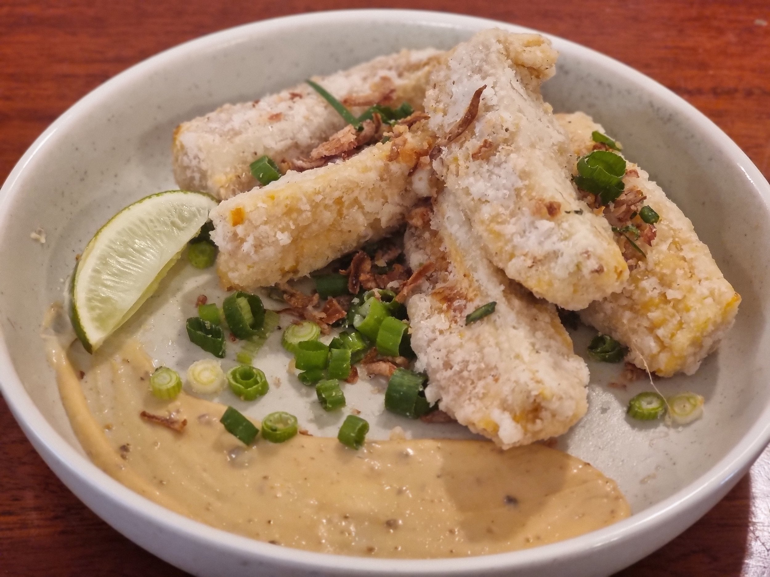 Bowl of fried fish fillets garnished with chopped green onions, served with a lemon wedge, accompanied by a creamy mustard sauce.