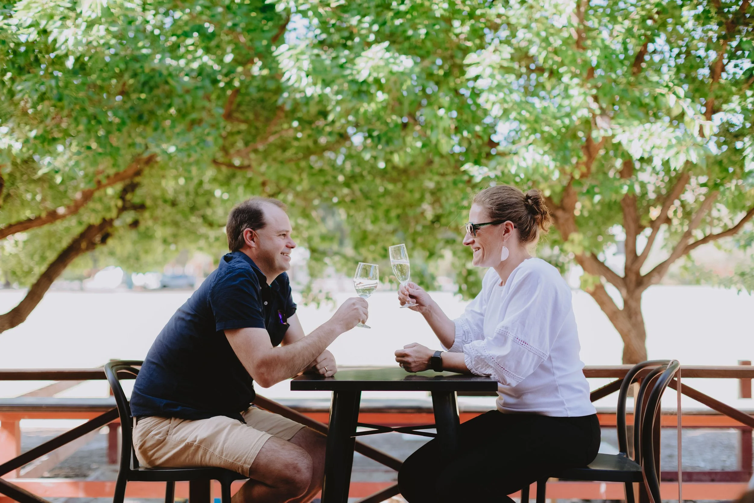 A man and woman sitting across from each other at a black outdoor table, smiling and holding glasses of white wine, with green trees in the background.