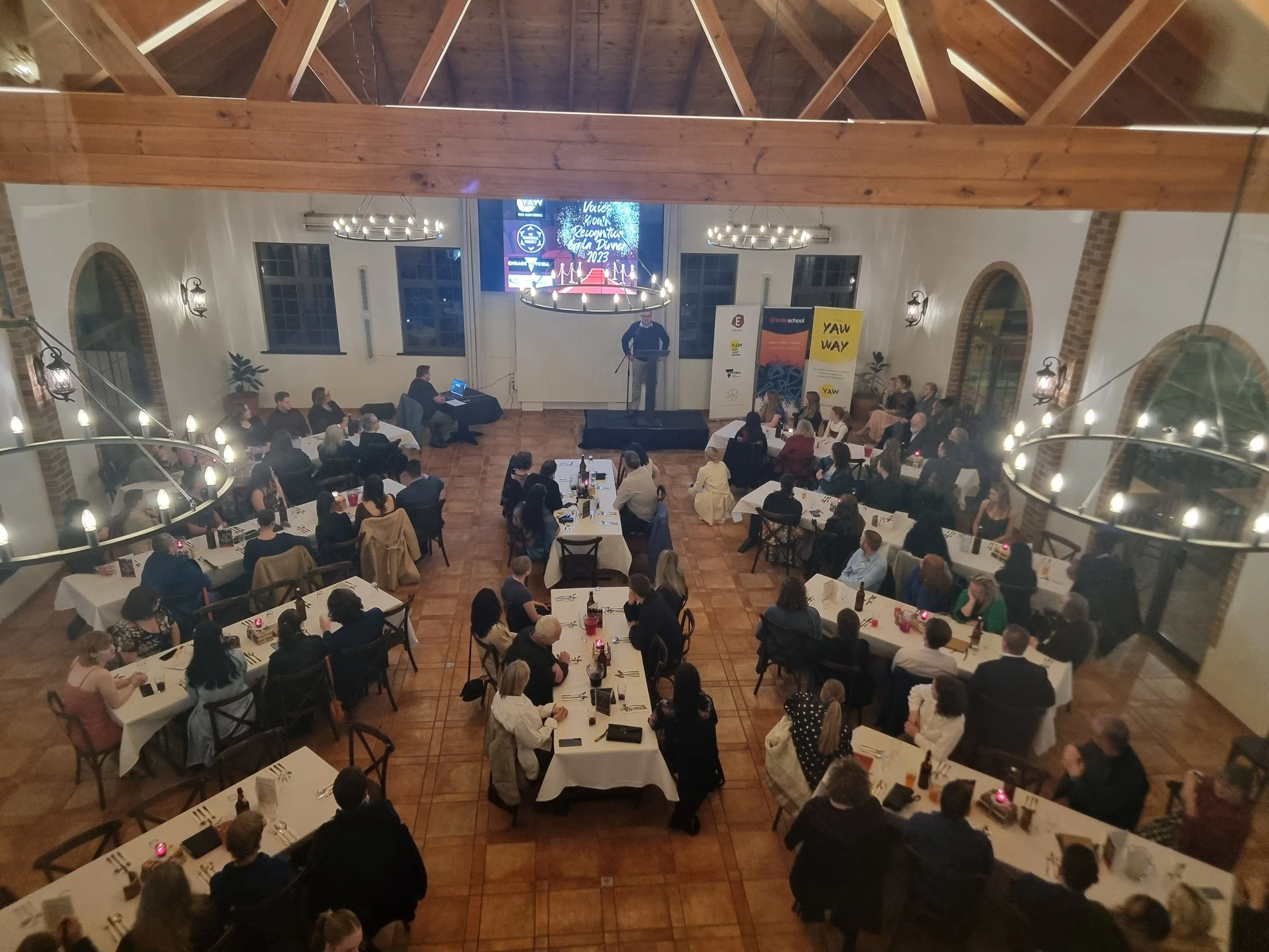 A large indoor banquet hall filled with people seated at tables, listening to a speaker on stage. The hall has high ceilings with exposed wooden beams, large windows, and elegant lighting fixtures, with banners and a screen displaying event informati