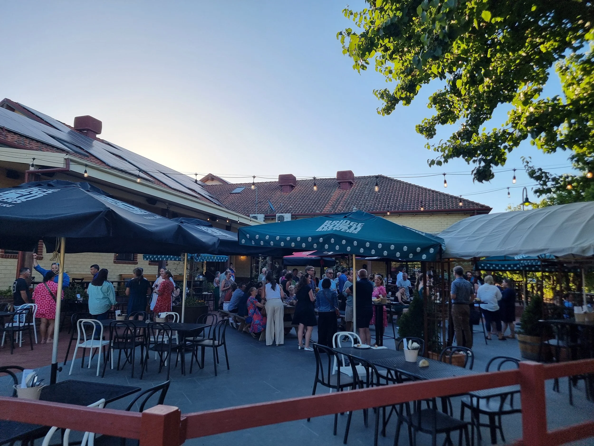 An outdoor gathering of people at a patio with umbrellas and string lights in the evening.