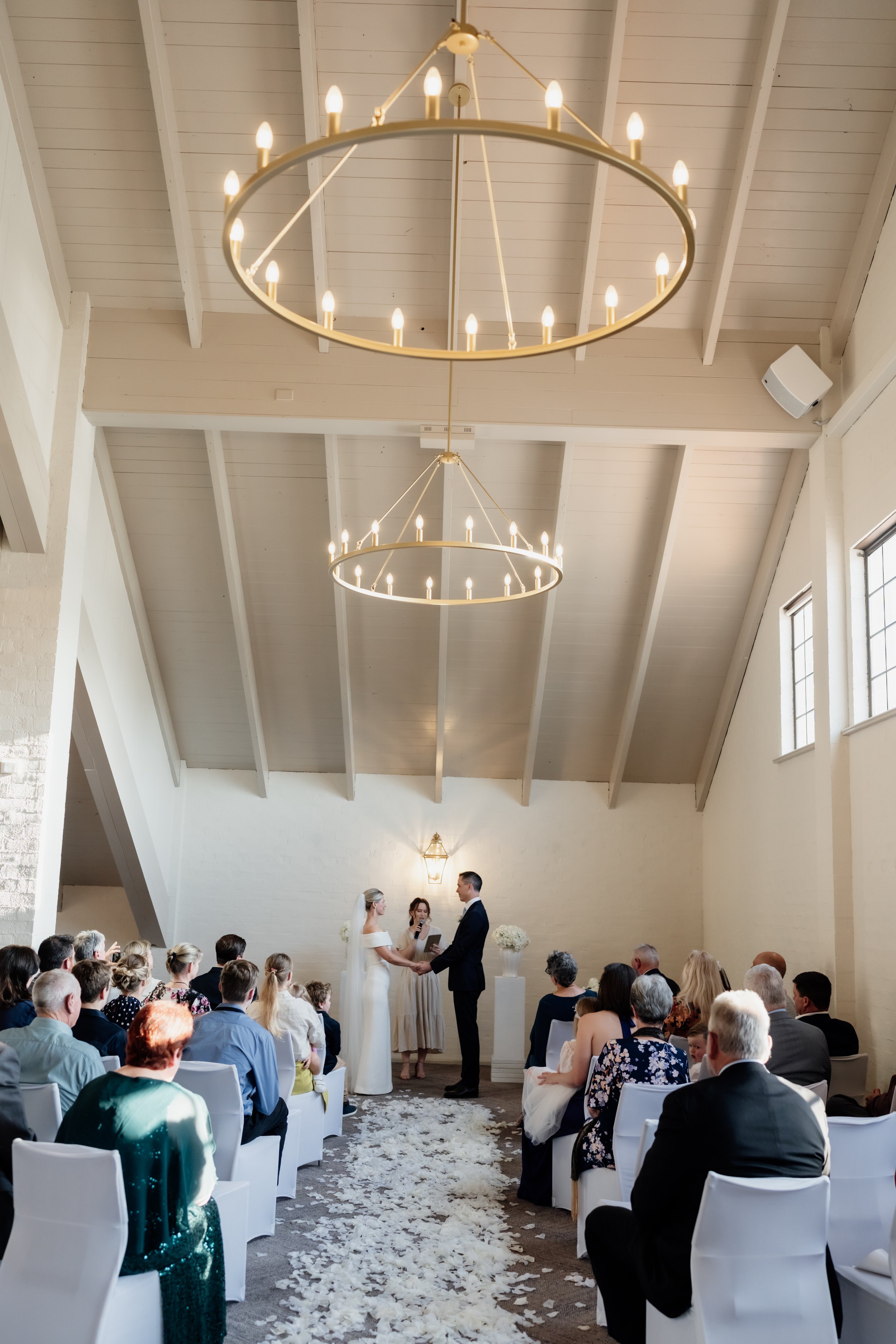 A wedding ceremony inside a bright, modern chapel with white walls and high ceilings, featuring hanging circular chandeliers, with a bride and groom holding hands and a officiant standing beside them, surrounded by seated guests.