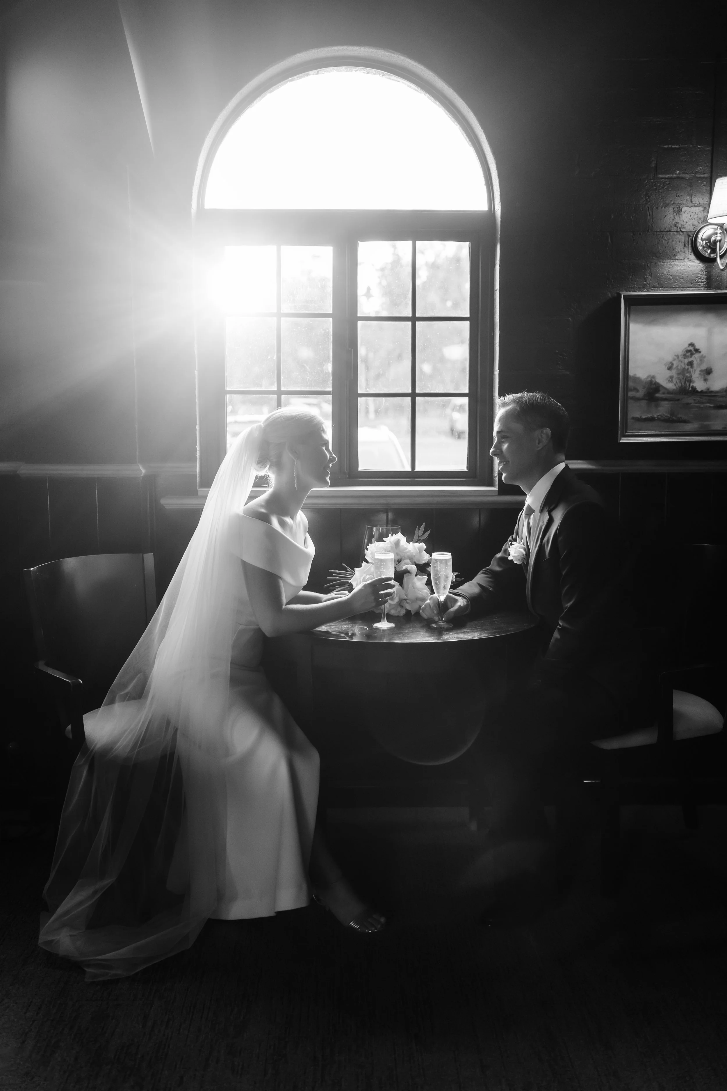 A black-and-white photo of a bride and groom sitting at a small round table with a flower arrangement, facing each other and holding champagne glasses, in a room with a large window and sunlight streaming in.