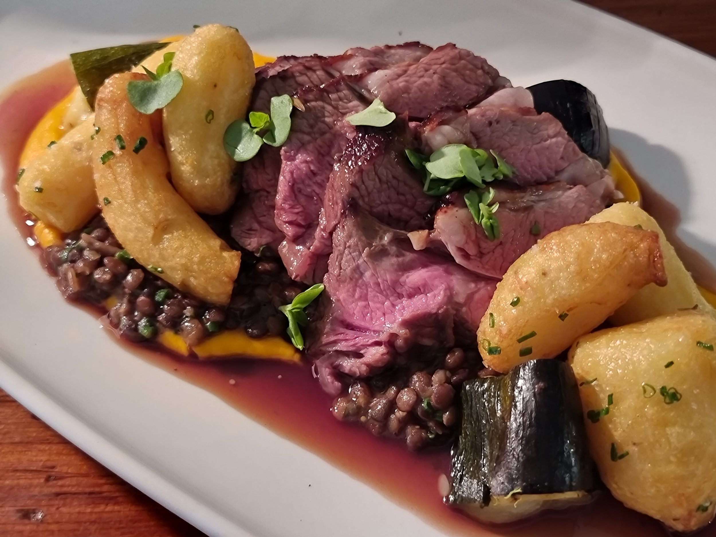 A plate of sliced medium-rare beef tenderloin garnished with microgreens, served with roasted potatoes, black lentils, and caramelized vegetables on a white plate.