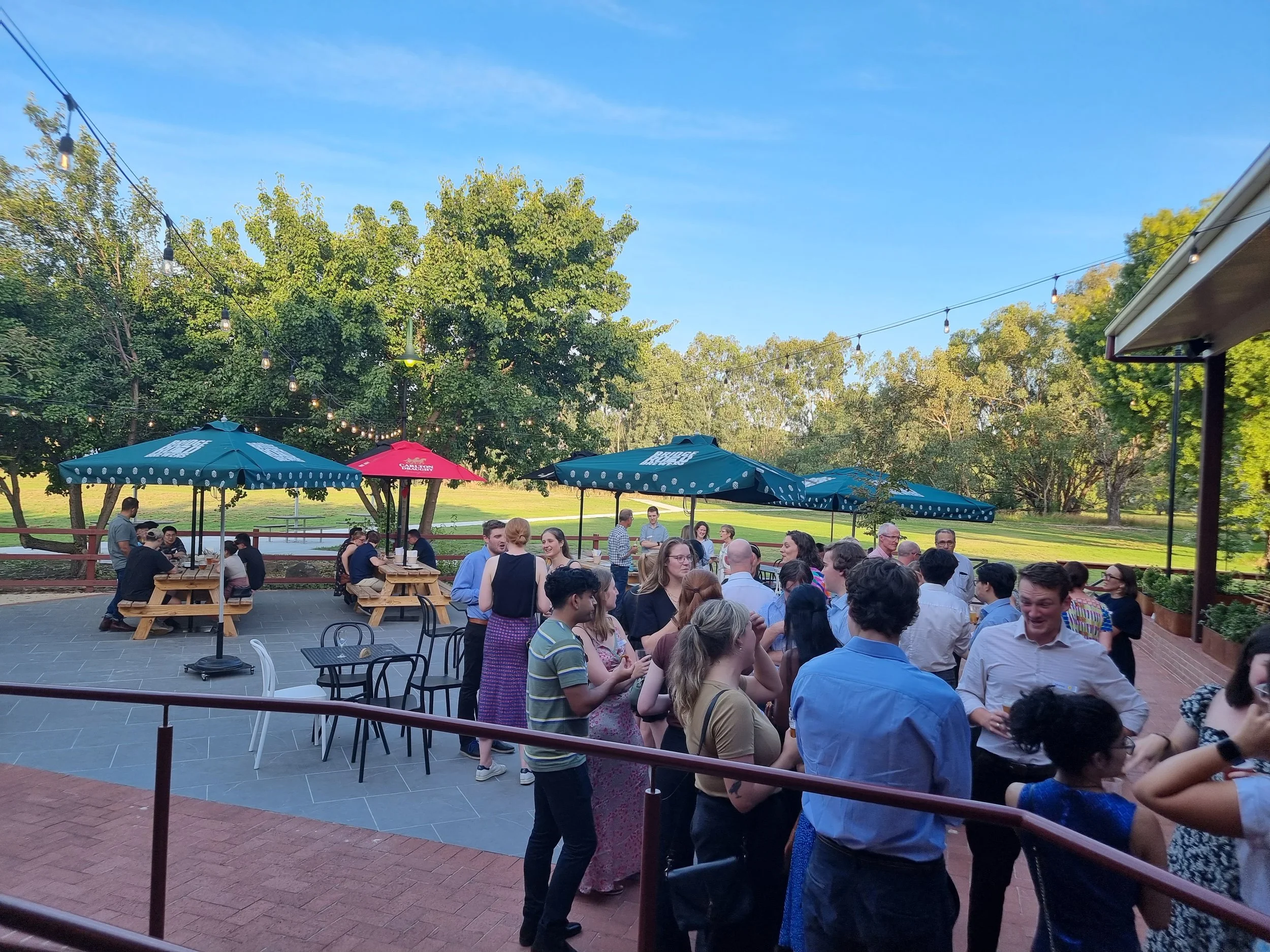People socializing outdoors at a gathering with umbrellas, picnic tables, and string lights on a sunny day.