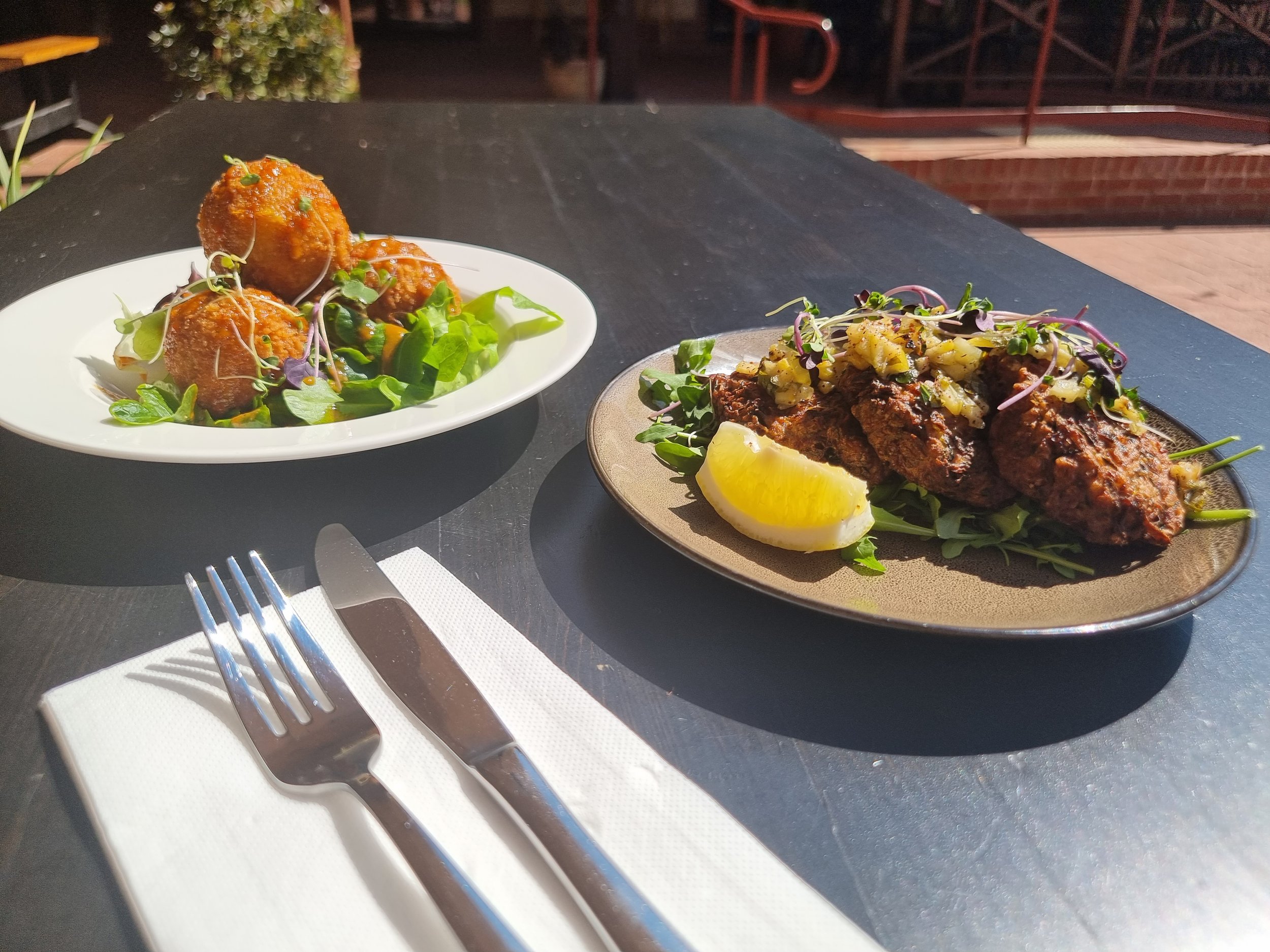Plate of three seasoned meatballs with microgreens on a white dish, and skewered grilled meat topped with chopped vegetables and microgreens, with a lemon wedge on a tan plate, on a black outdoor table.