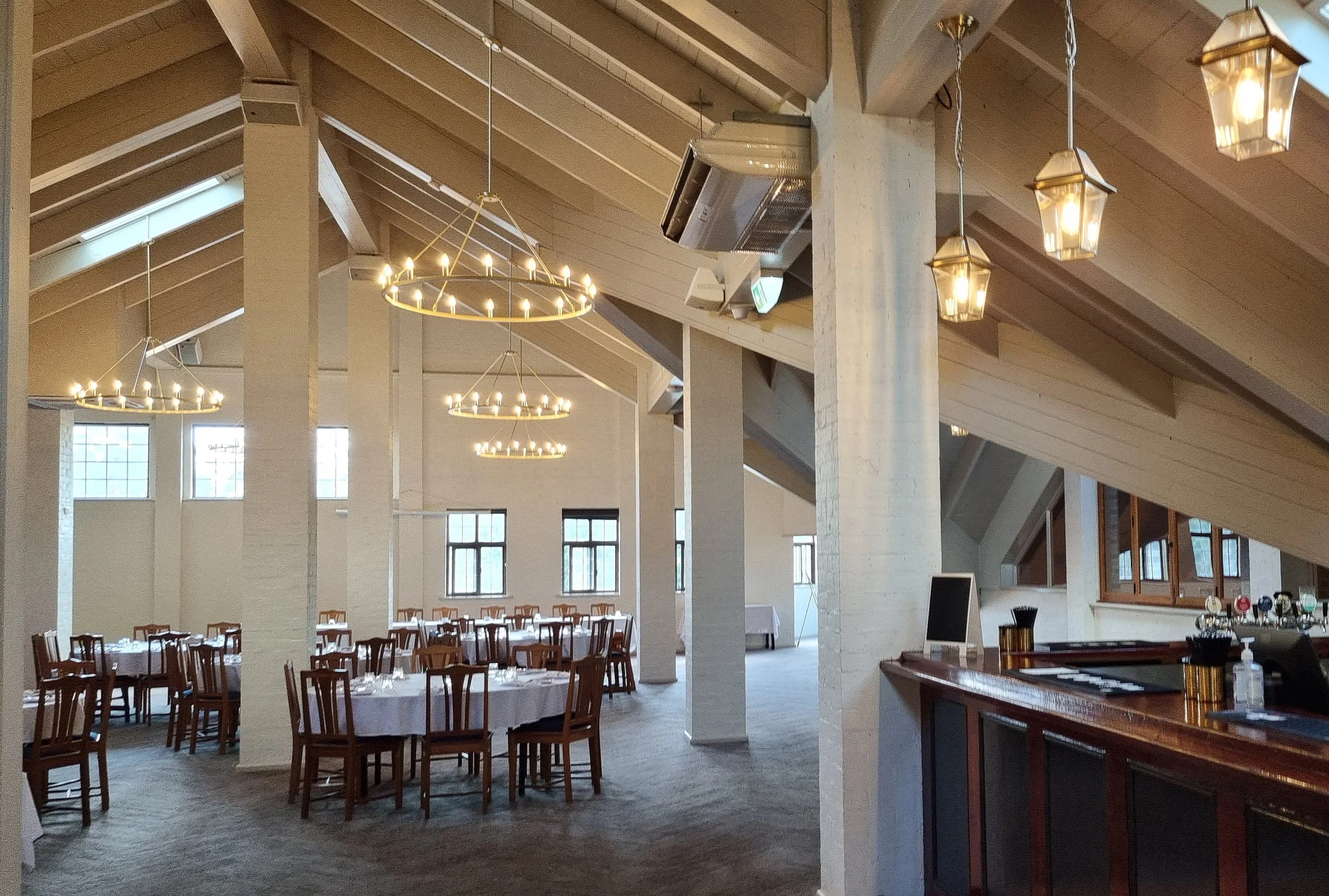 Dining area with round tables and wooden chairs, decorated for an event, with chandeliers and pendant lights hanging from a sloped ceiling with large windows on the walls.